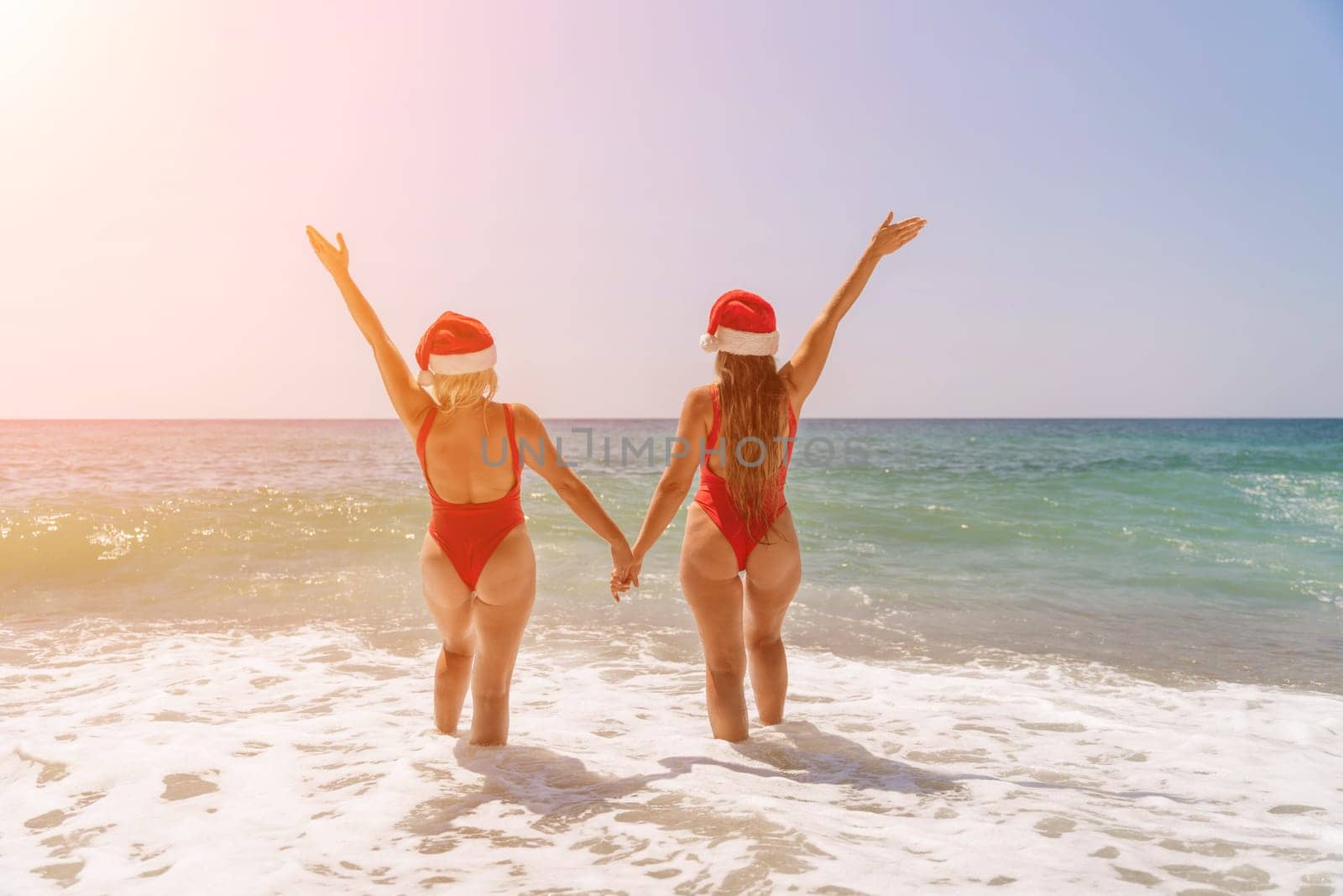 Women Santa hats ocean play. Seaside, beach daytime, enjoying beach fun. Two women in red swimsuits and Santa hats are enjoying themselves in the ocean waves and raising their hands up. by Matiunina