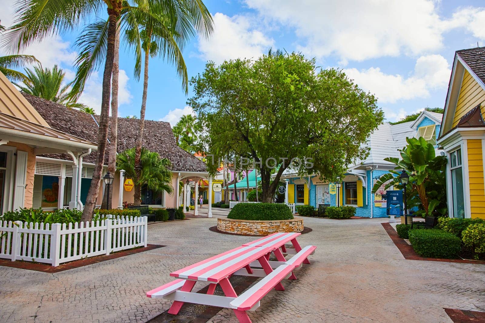 Vibrant tropical shopping village in Nassau, Bahamas, featuring pastel colored buildings, a pink picnic table, and lush palm trees.