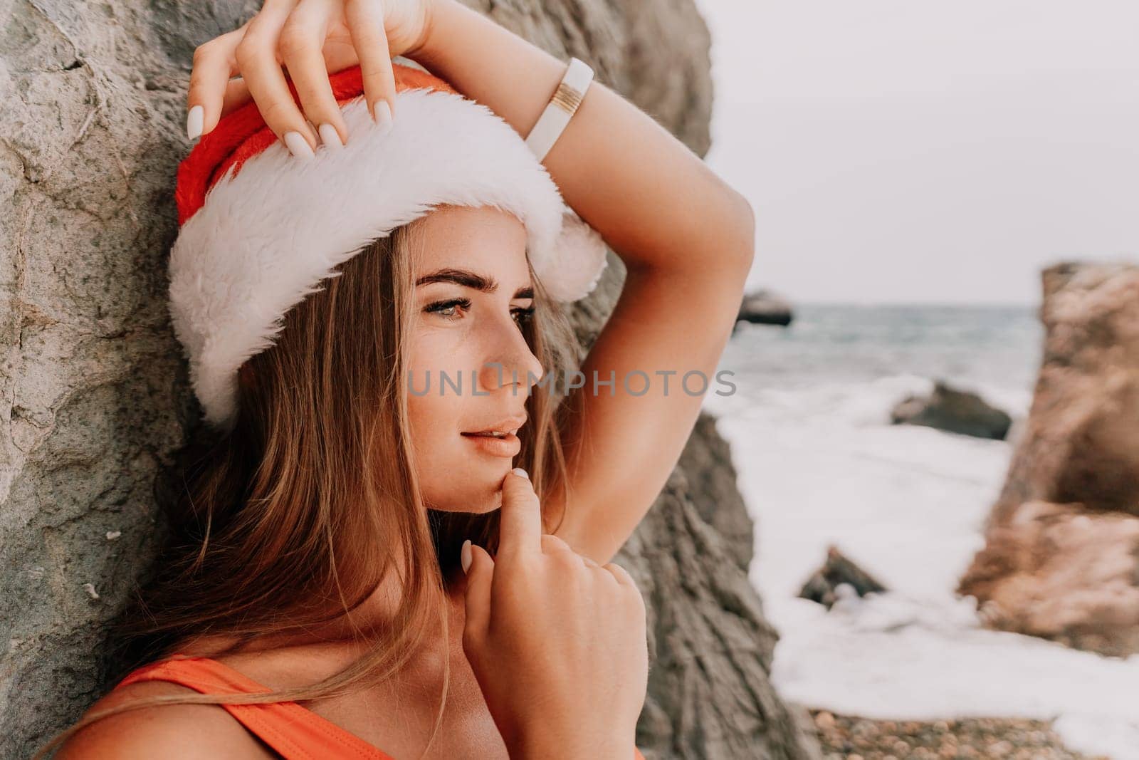 Woman summer travel sea. Happy tourist in red bikini and Santas hat enjoy taking picture outdoors for memories. Woman traveler posing on the beach surrounded by volcanic mountains, sharing travel joy by panophotograph