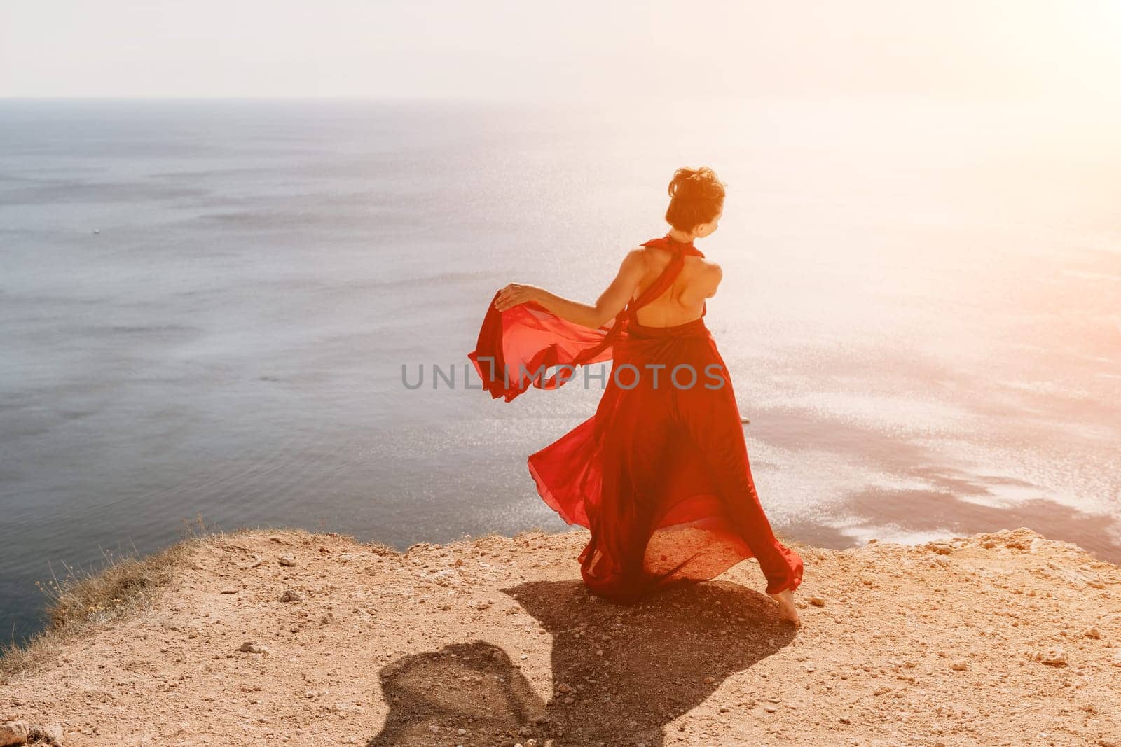 Side view a Young beautiful sensual woman in a red long dress posing on a rock high above the sea during sunrise. Girl on the nature on blue sky background. Fashion photo.