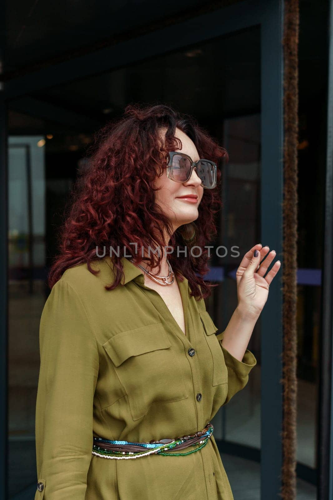 Woman leaves a supermarket. Caucasian model with long brunette hair, wears sunglasses and a khaki dress. by Matiunina