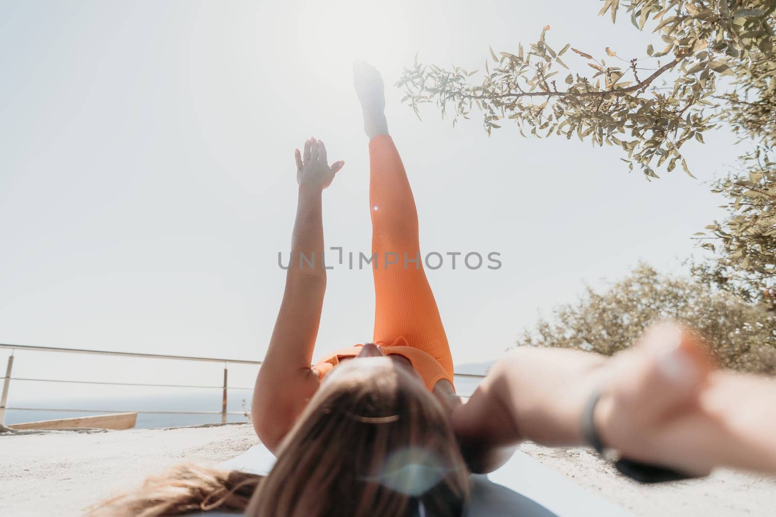 Fitness woman sea. Happy middle aged woman in orange sportswear exercises morning outdoors on yoga mat with laptop in park over ocean beach. Female fitness pilates yoga routine. Healthy lifestyle. by panophotograph