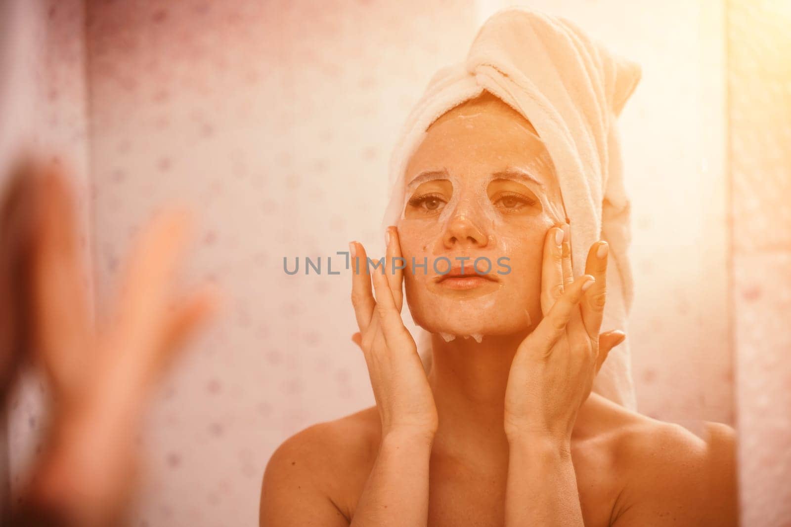 Young beautiful woman using a moisturizing facial mask after taking a bath. Pretty attractive girl in a towel on her head stands in front of a mirror in a home bathroom. Daily hygiene and skin care by Matiunina