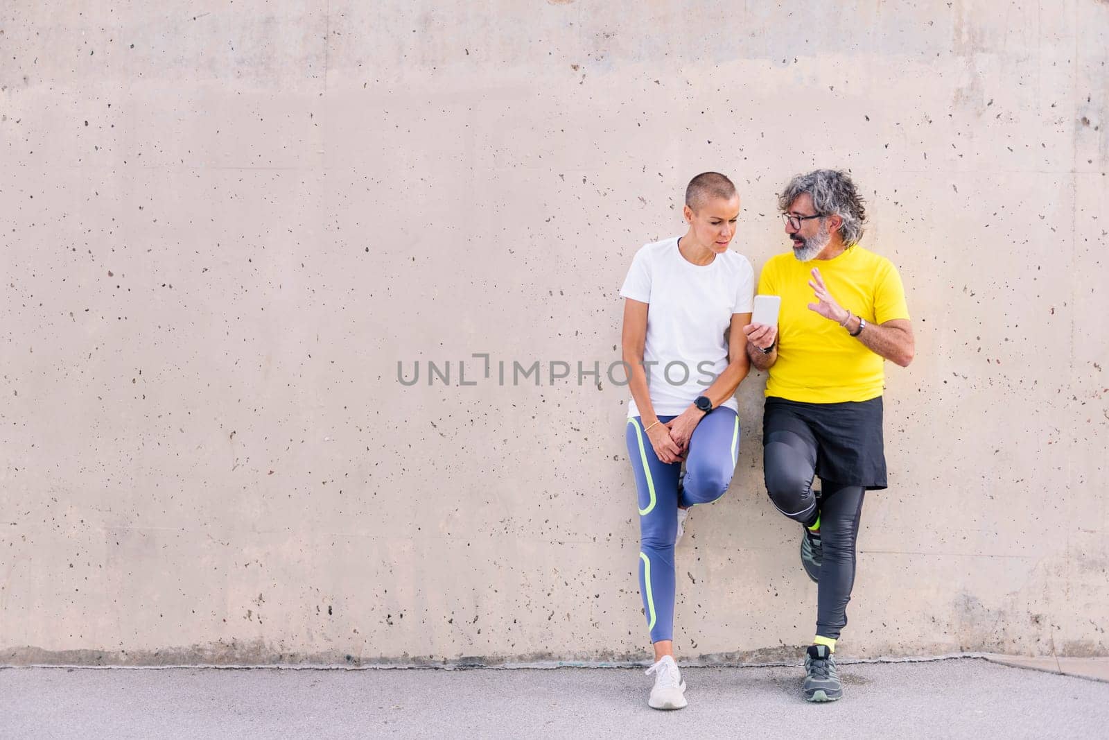 senior sports man and young sports woman talking and using mobile phone leaning an a wall, concept of technology of communication, copy space for text