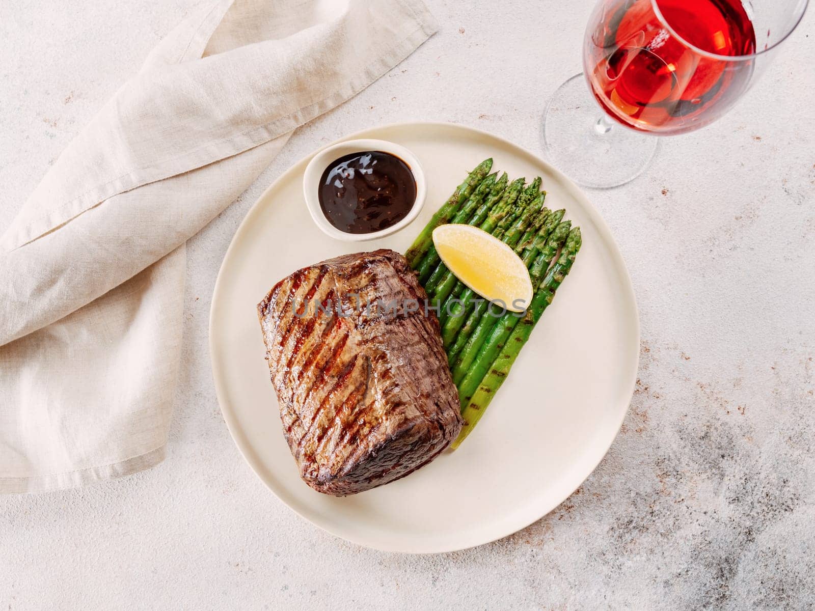 Grilled meat steak on plate with lemon and asparagus on white table background. Food and cuisine concept. Restaurant-style plating, with red wine glass on table. Top view or flat lay