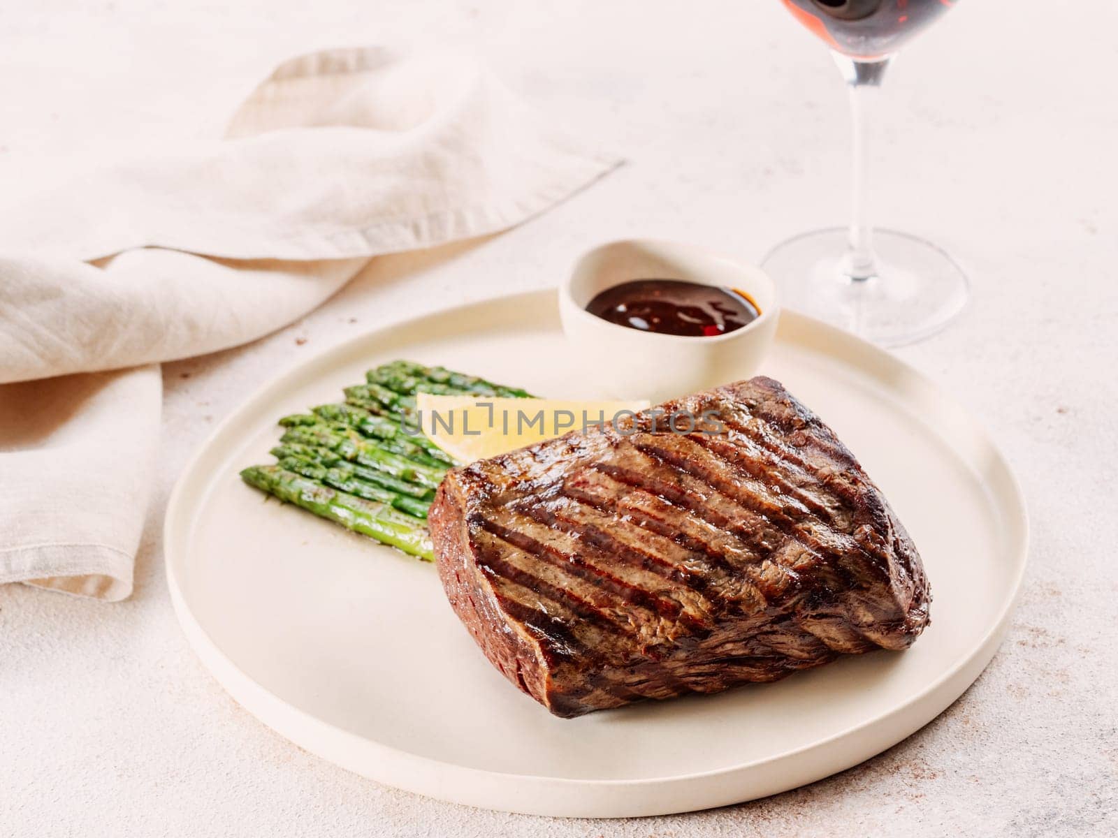 Grilled meat steak on plate with lemon and asparagus on white table background. Food and cuisine concept. Restaurant-style plating, with red wine glass on table