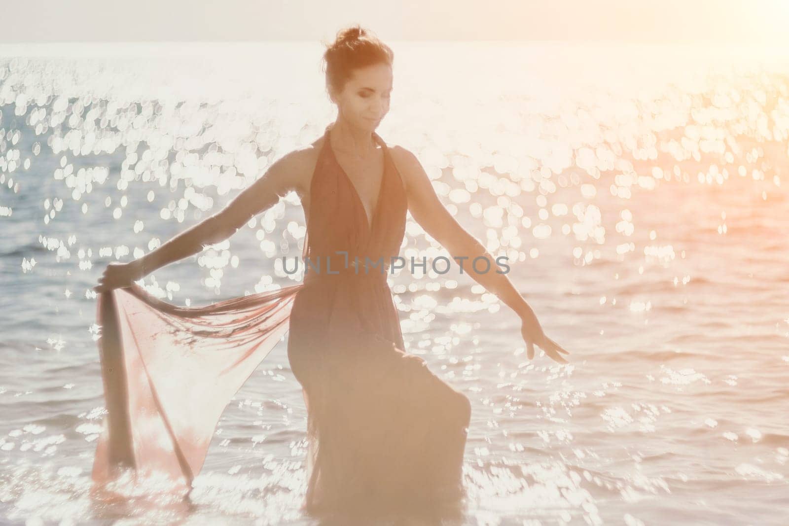 Side view a Young beautiful sensual woman in a red long dress posing on a rock high above the sea during sunrise. Girl on the nature on blue sky background. Fashion photo.