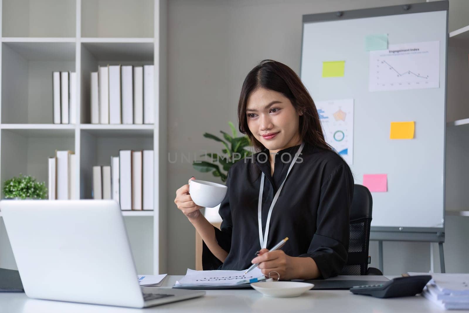 Beautiful smiling Asian businesswoman working using laptop and drinking coffee Asian businesswoman working with finance and calculator in her office by wichayada