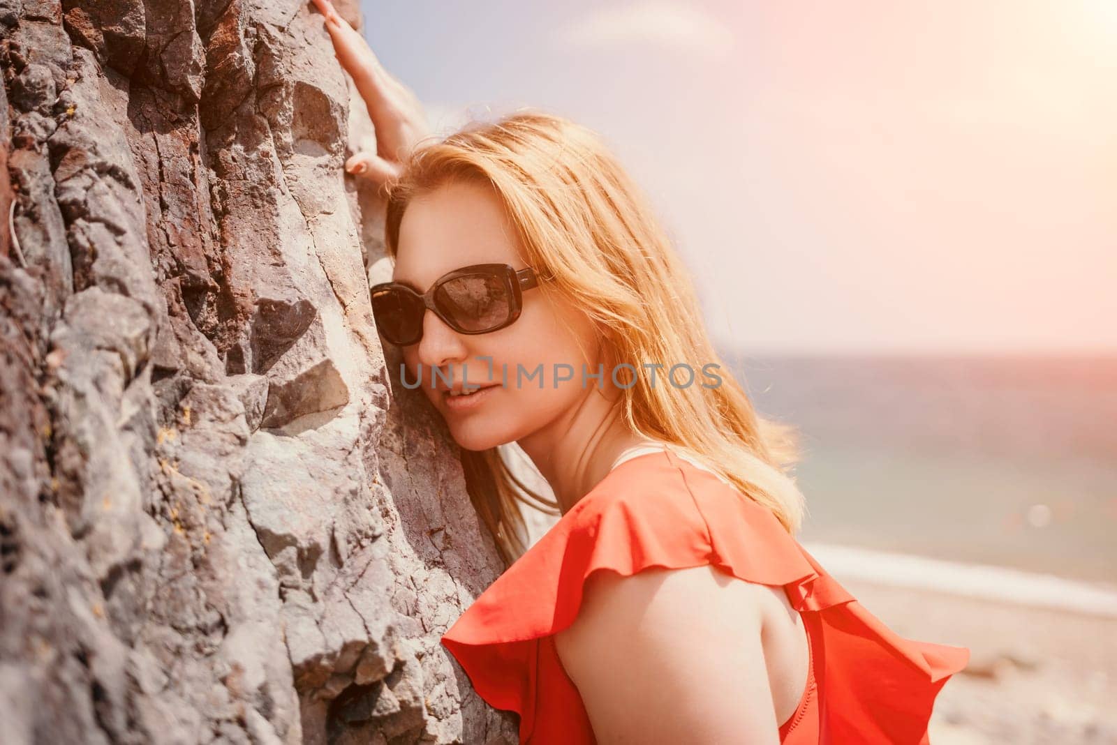 Young woman in red bikini on Beach. Blonde in sunglasses on pebble beach enjoying sun. Happy lady in one piece red swimsuit relaxing and sunbathing by turquoise sea ocean on hot summer day. Close up by panophotograph