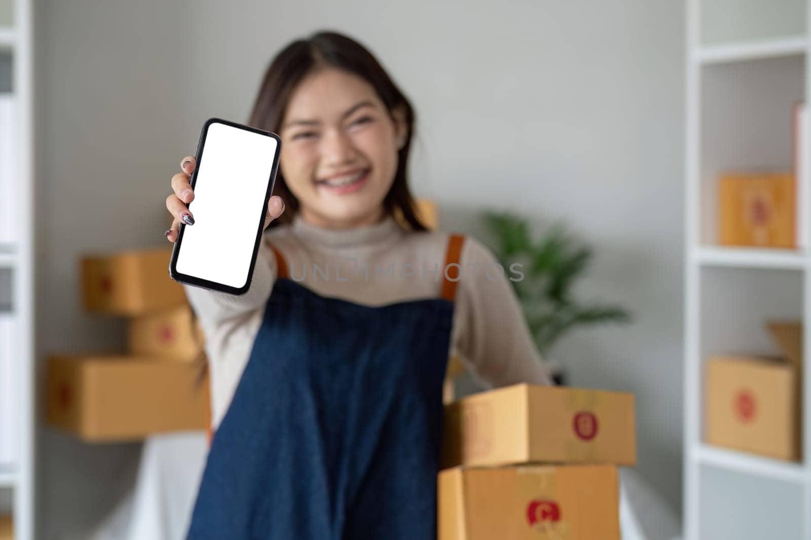 Beautiful smiling Asian woman holding a cardboard box and hands showing a smartphone mockup of a blank screen. Advertise your screen Online delivery concept by wichayada