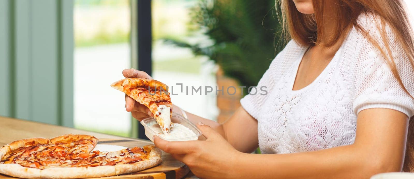 A girl dips a slice of pizza in sauce in a plastic container before eating it