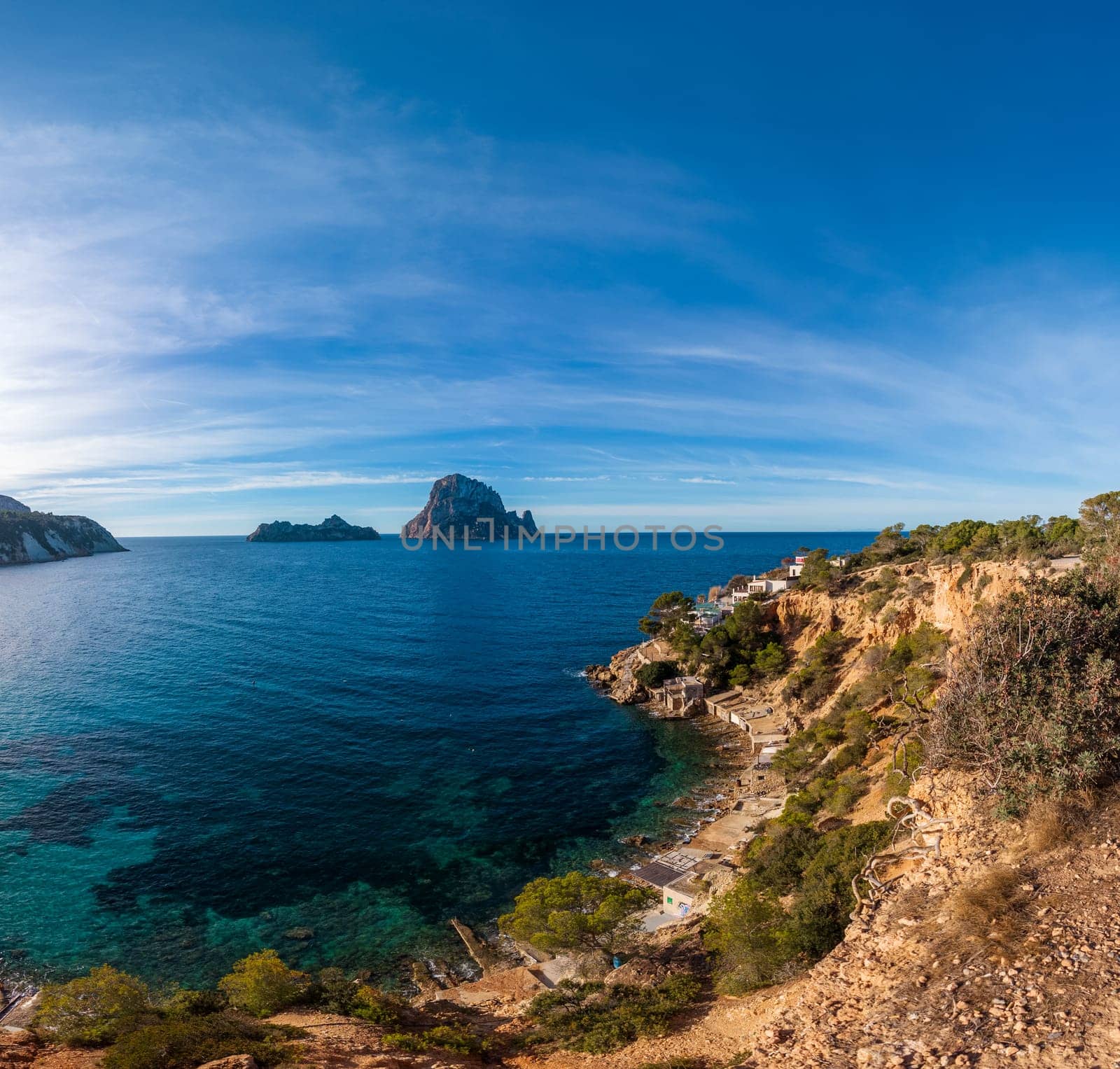 Picturesque coastal view with rocky formations and cliff-edge structures under a bright blue sky, Es Vedra in Ibiza