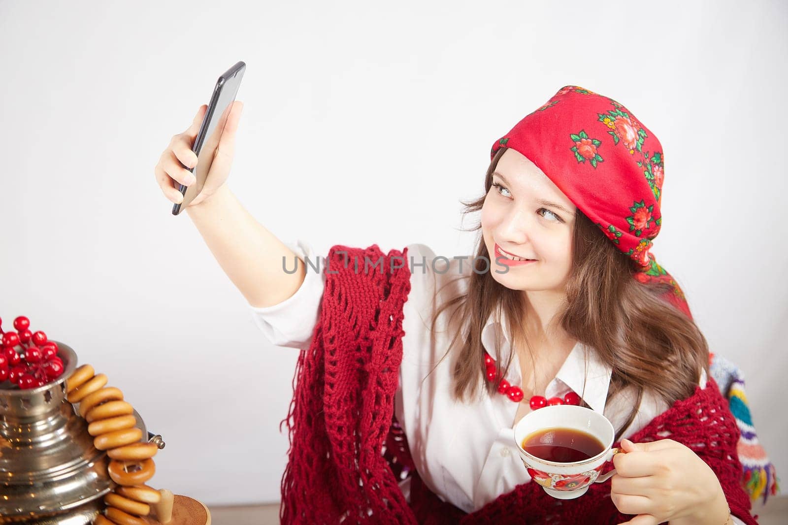 A fashionable modern girl in stylized folk clothes at a table with a samovar, bagels and tea takes a selfie on the Orthodox holiday of Maslenitsa and Easter. Funny photo shoot for a young woman by keleny