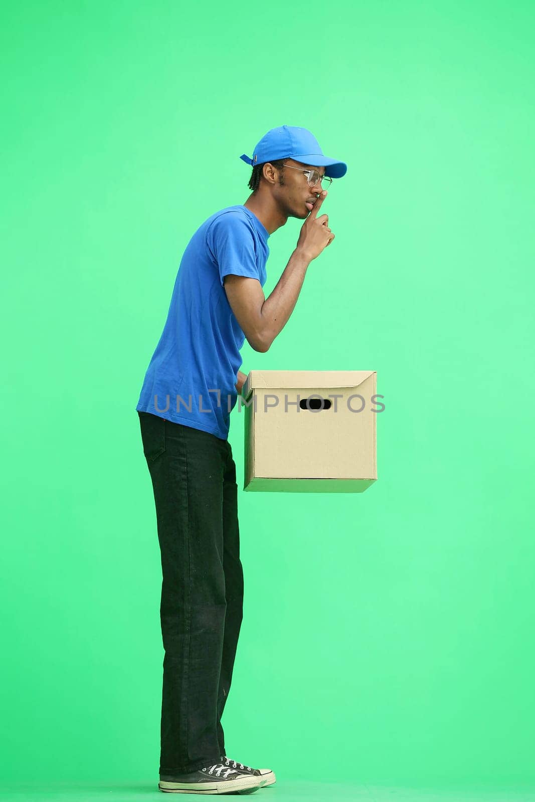 A male deliveryman, on a green background, in full height, with a box, shows a sign of silence by Prosto