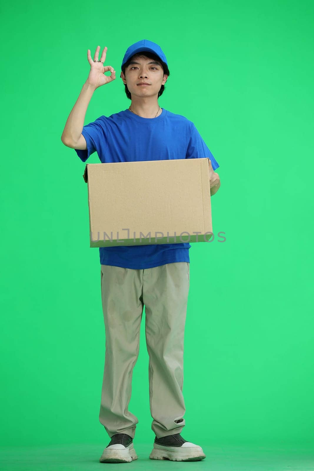 The delivery guy, on a green background, full-length, with a box, shows the ok sign.