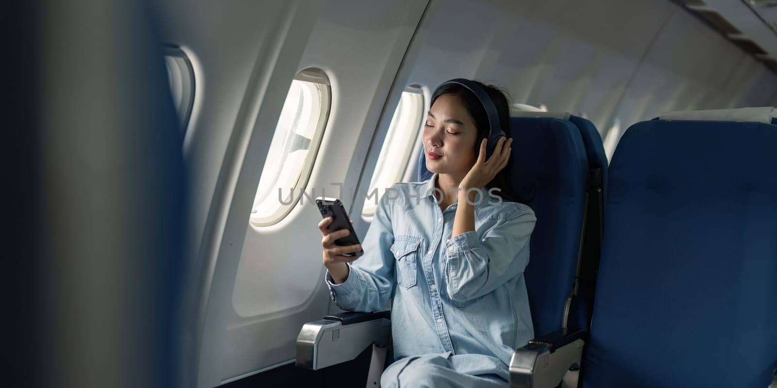 Asian woman traveler in airplane wearing headset listening music from mobile phone going on a trip vacation travel concept.