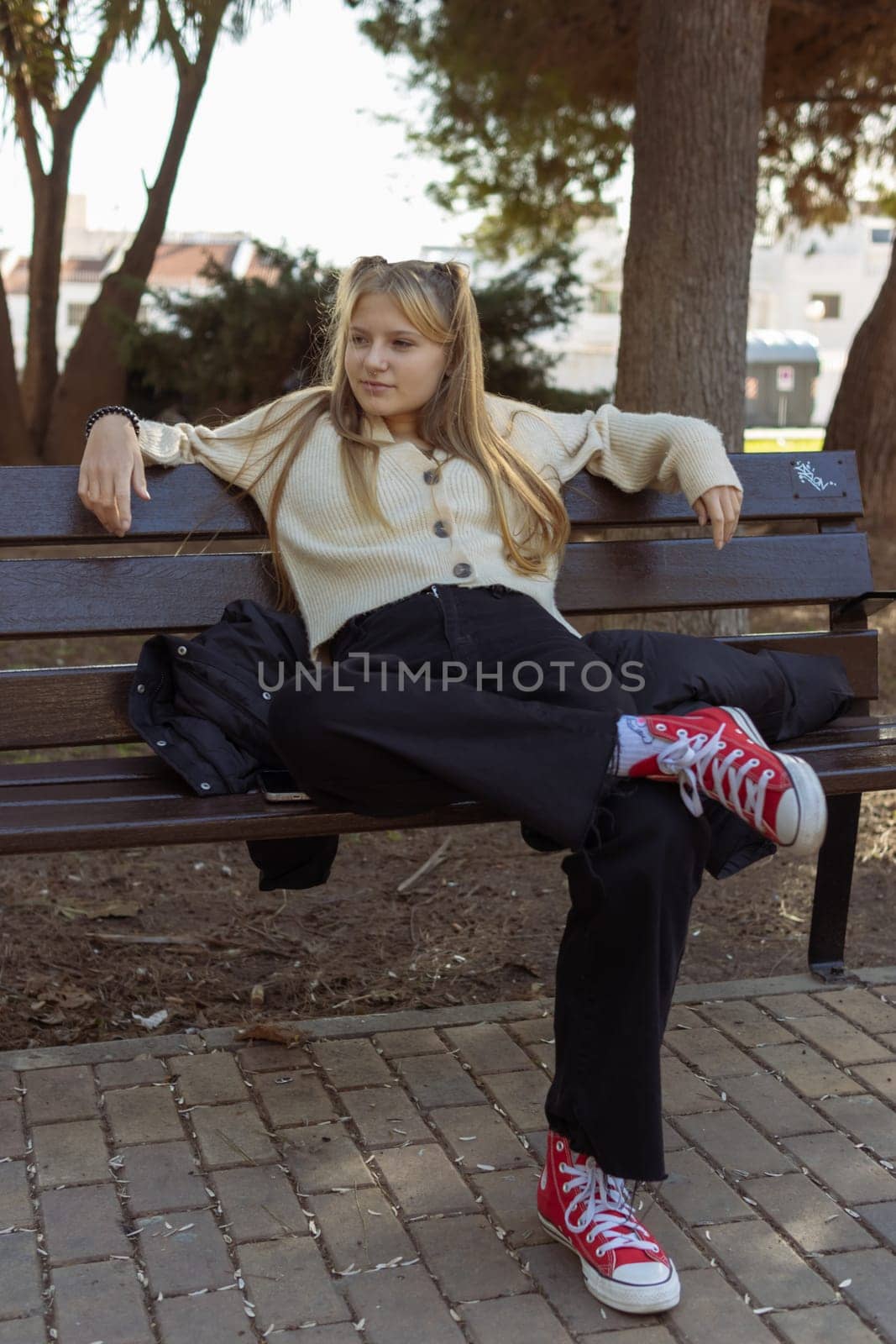 vertical photo a girl of fair appearance with a piercing in the nose dressed in a white jacket, black jeans and red sneakers sits on a bench, close-up on a hipster girl. High quality photo