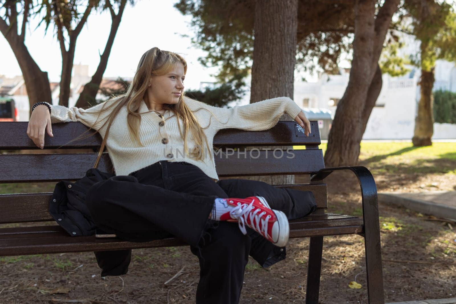 a teenage girl of fair appearance with a piercing in the nose dressed in a white jacket, black jeans and red sneakers sits on a bench, close-up on a hipster girl. High quality photo