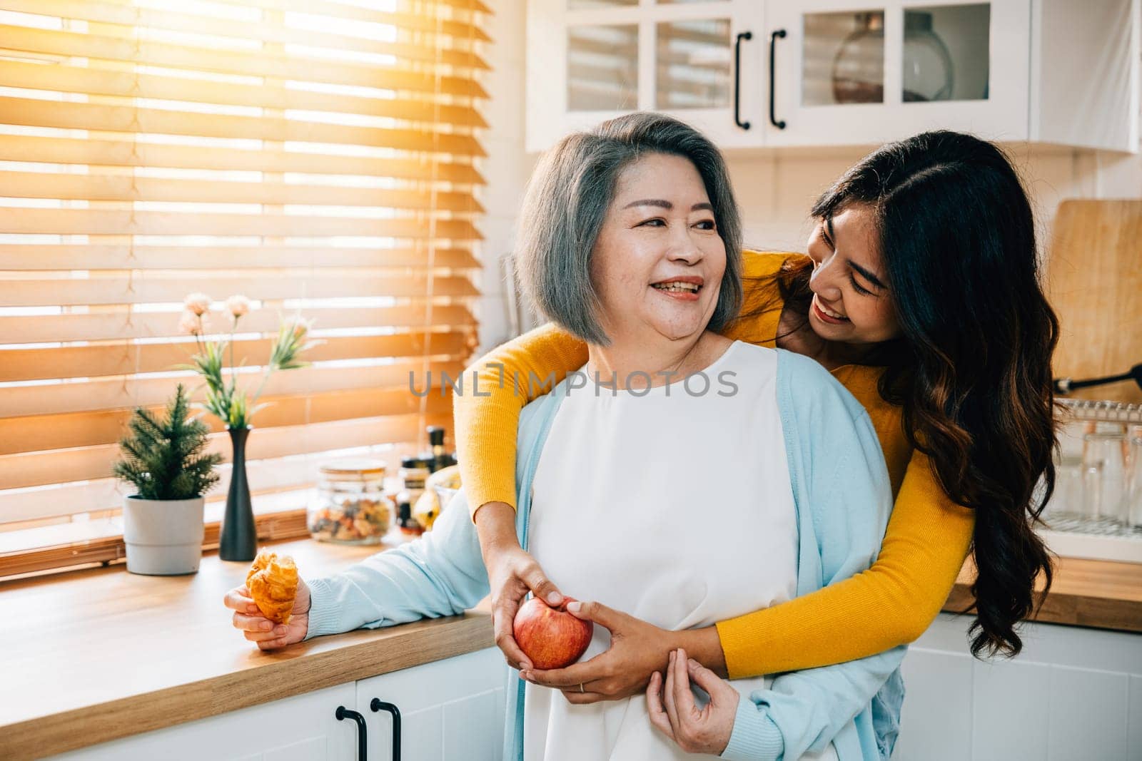 A smiling old woman stands in the kitchen, her adult daughter, a cheerful young woman in an apron, holds an apple. Their bond exemplifies the joy of teaching, learning, and family togetherness. by Sorapop