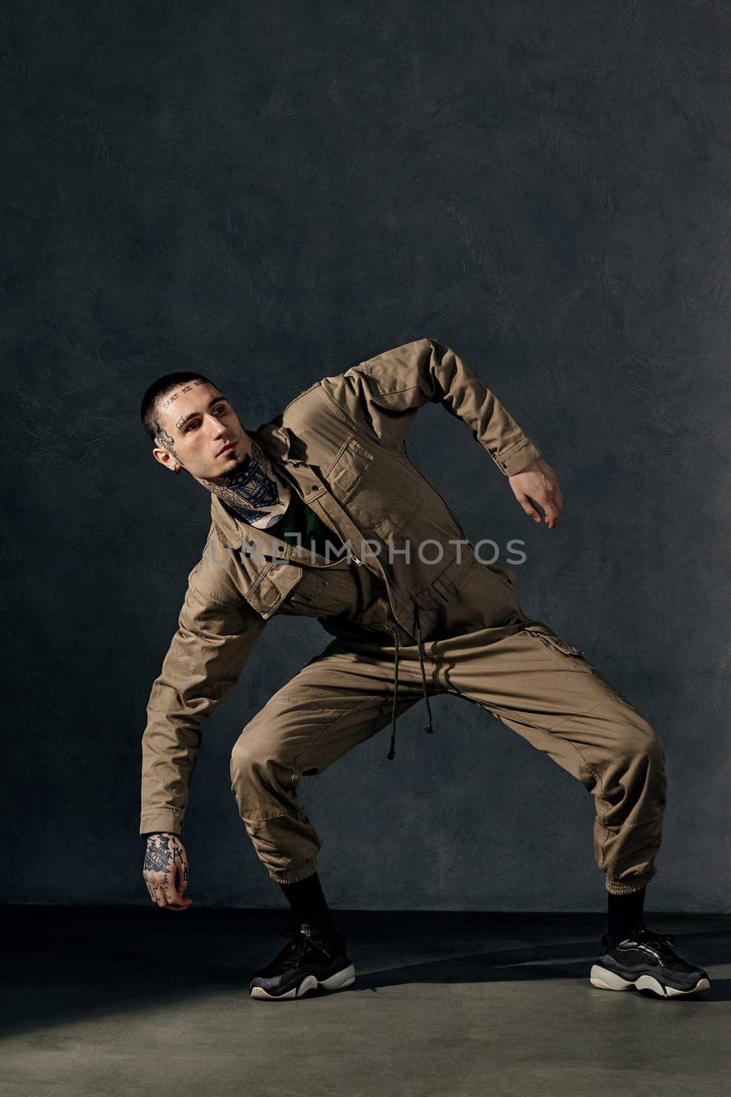 Young guy with tattooed body and face, earrings, beard. Dressed in khaki overalls and black sneakers. Dancing on gray background. Dancehall, hip-hop by nazarovsergey