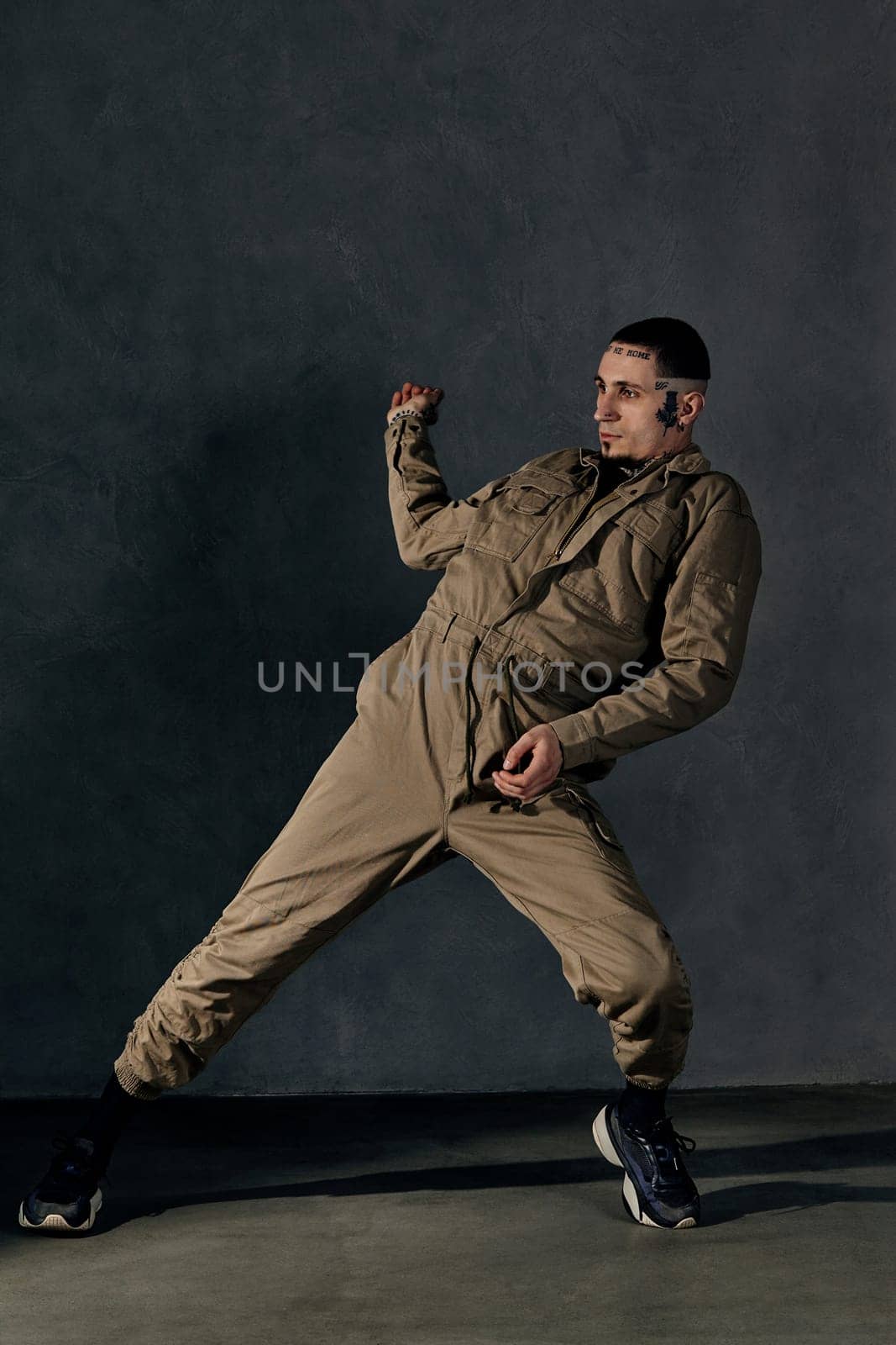 Young stylish performer with tattooed body and face, earrings, beard. Dressed in khaki overalls and black sneakers. He is dancing against gray background. Dancehall, hip-hop. Full length, copy space