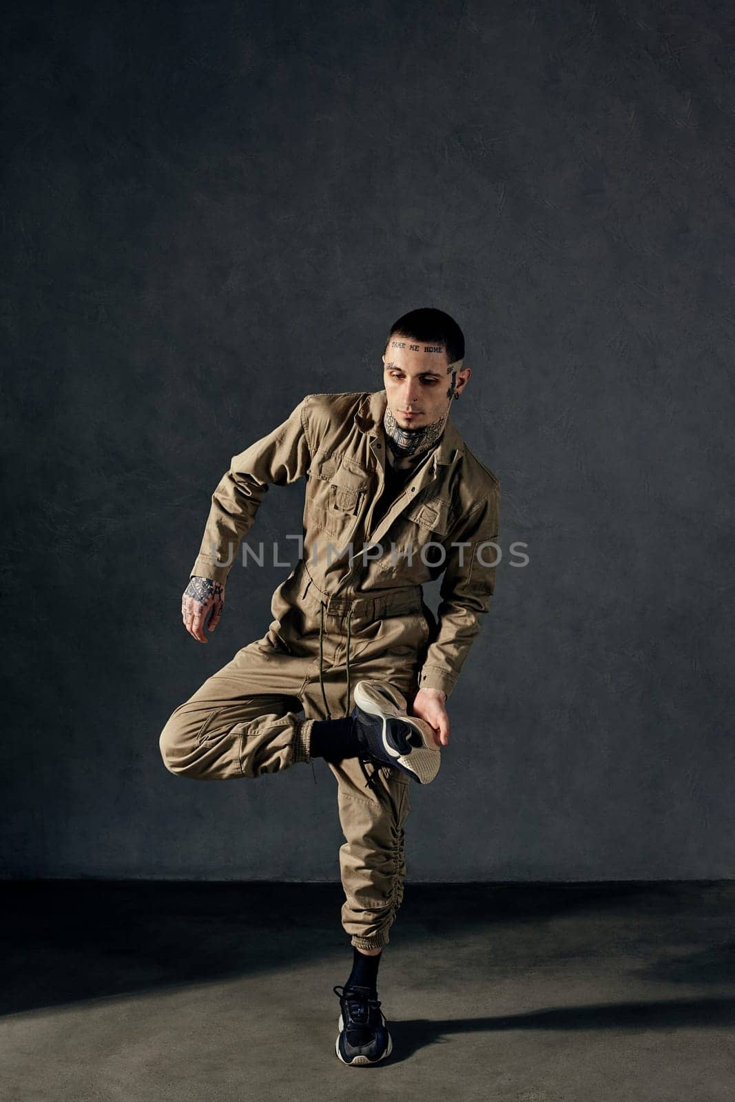 Young handsome man with tattooed body and face, earrings, beard. Dressed in khaki overalls and black sneakers. He is dancing against gray studio background. Dancehall, hip-hop. Full length, copy space
