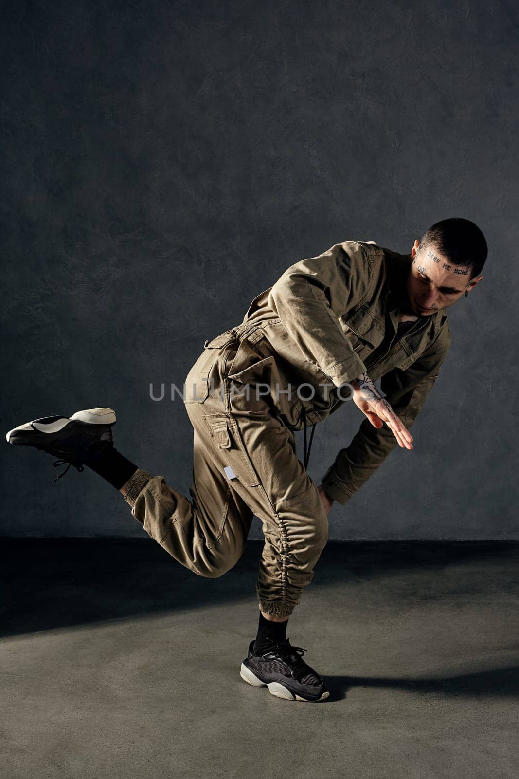 Young handsome fellow with tattooed body and face, earrings, beard. Dressed in khaki jumpsuit and black sneakers. He dancing against gray studio background. Dancehall, hip-hop. Full length, copy space