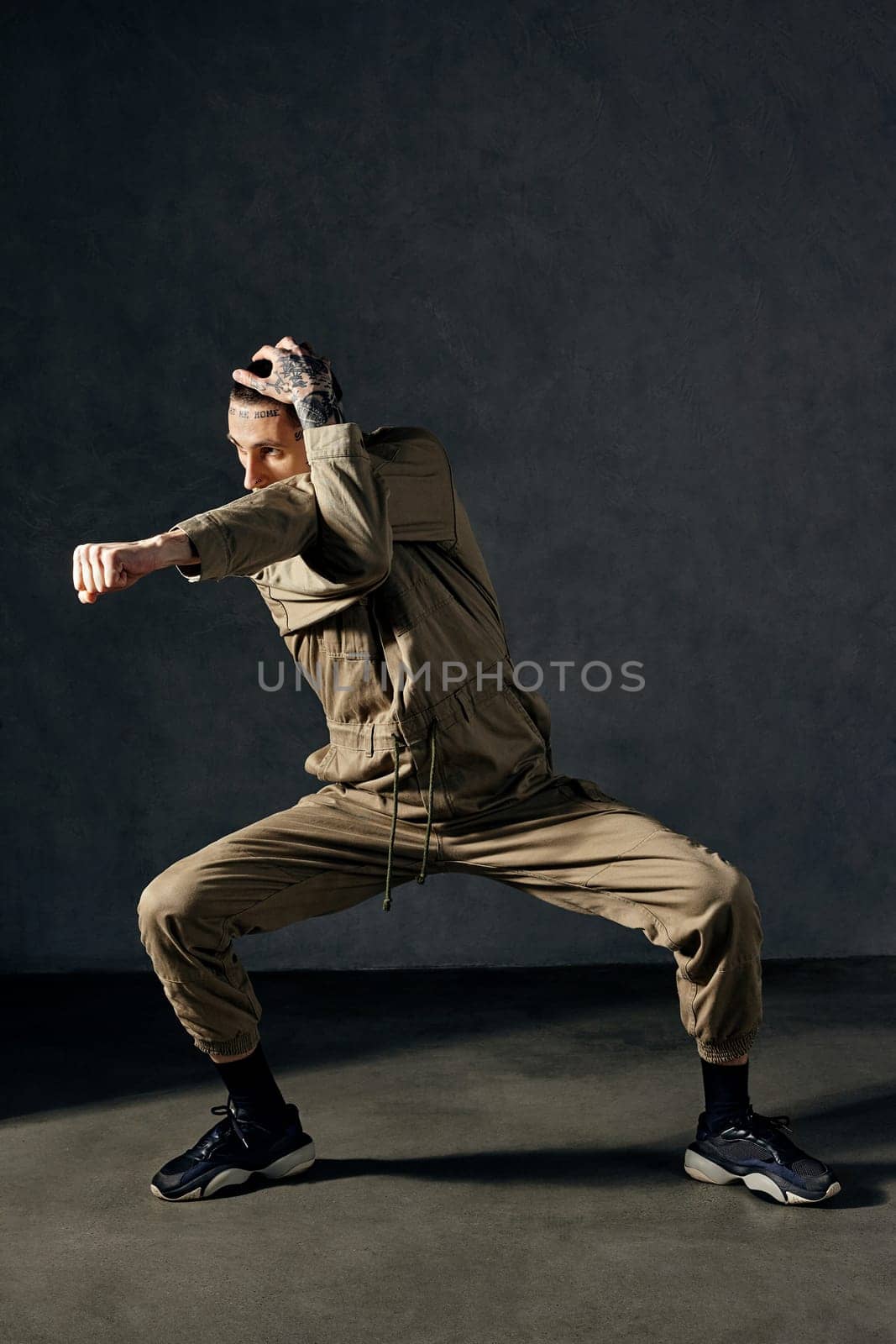Young graceful man with tattooed body and face, earrings, beard. Dressed in khaki overalls and black sneakers. He is dancing against gray studio background. Dancehall, hip-hop. Full length, copy space