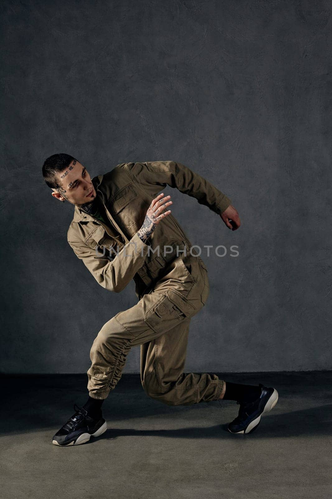 Graceful male with tattooed body and face, earrings, beard. Dressed in khaki jumpsuit, black sneakers. Dancing on gray background. Dancehall, hip-hop by nazarovsergey