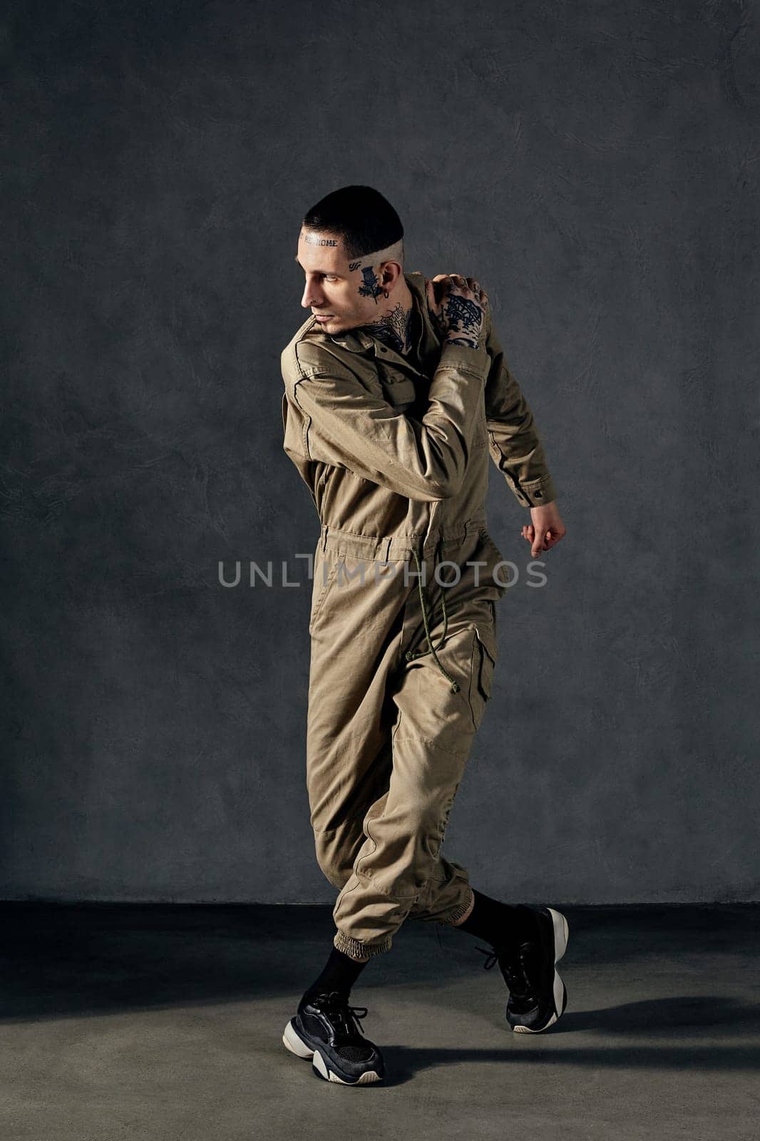 Young graceful fellow with tattooed body and face, earrings, beard. Dressed in khaki jumpsuit and black sneakers. He dancing against gray studio background. Dancehall, hip-hop. Full length, copy space