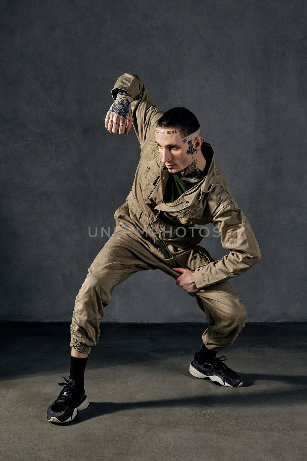 Modern fellow with tattooed body and face, earrings, beard. Dressed in khaki jumpsuit, black sneakers. Dancing on gray background. Dancehall, hip-hop by nazarovsergey