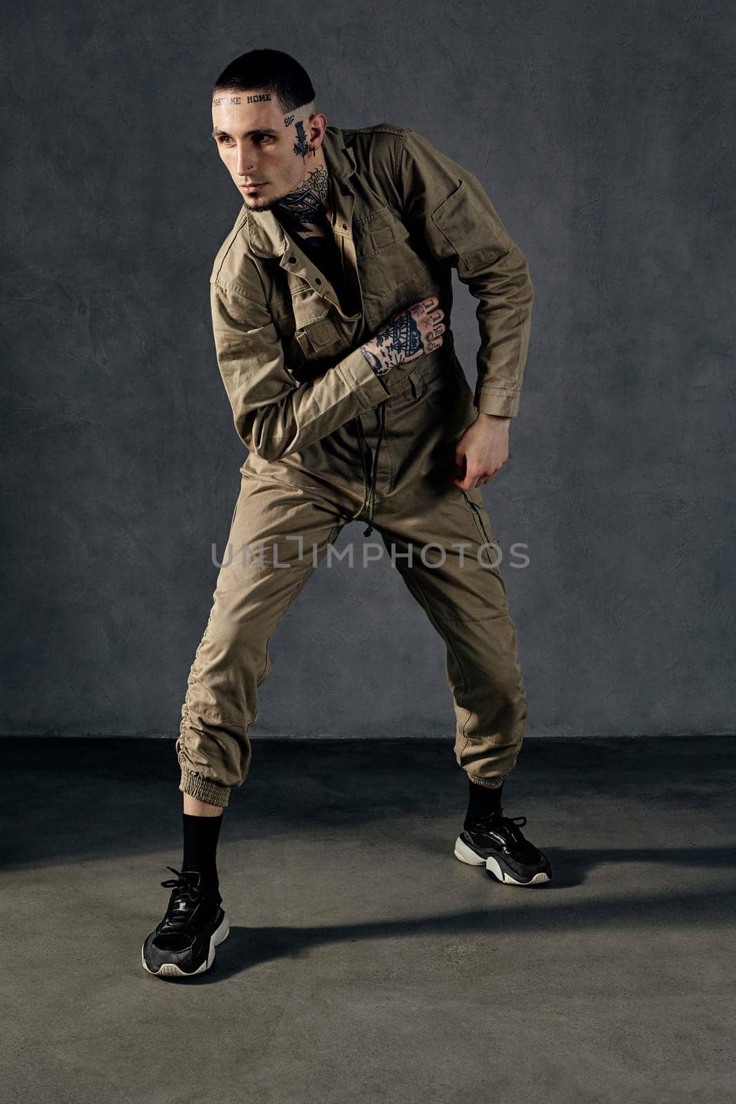 Modern performer with tattooed body, earrings, beard. Dressed in khaki overalls and black sneakers. Dancing on gray background. Dancehall, hip-hop by nazarovsergey