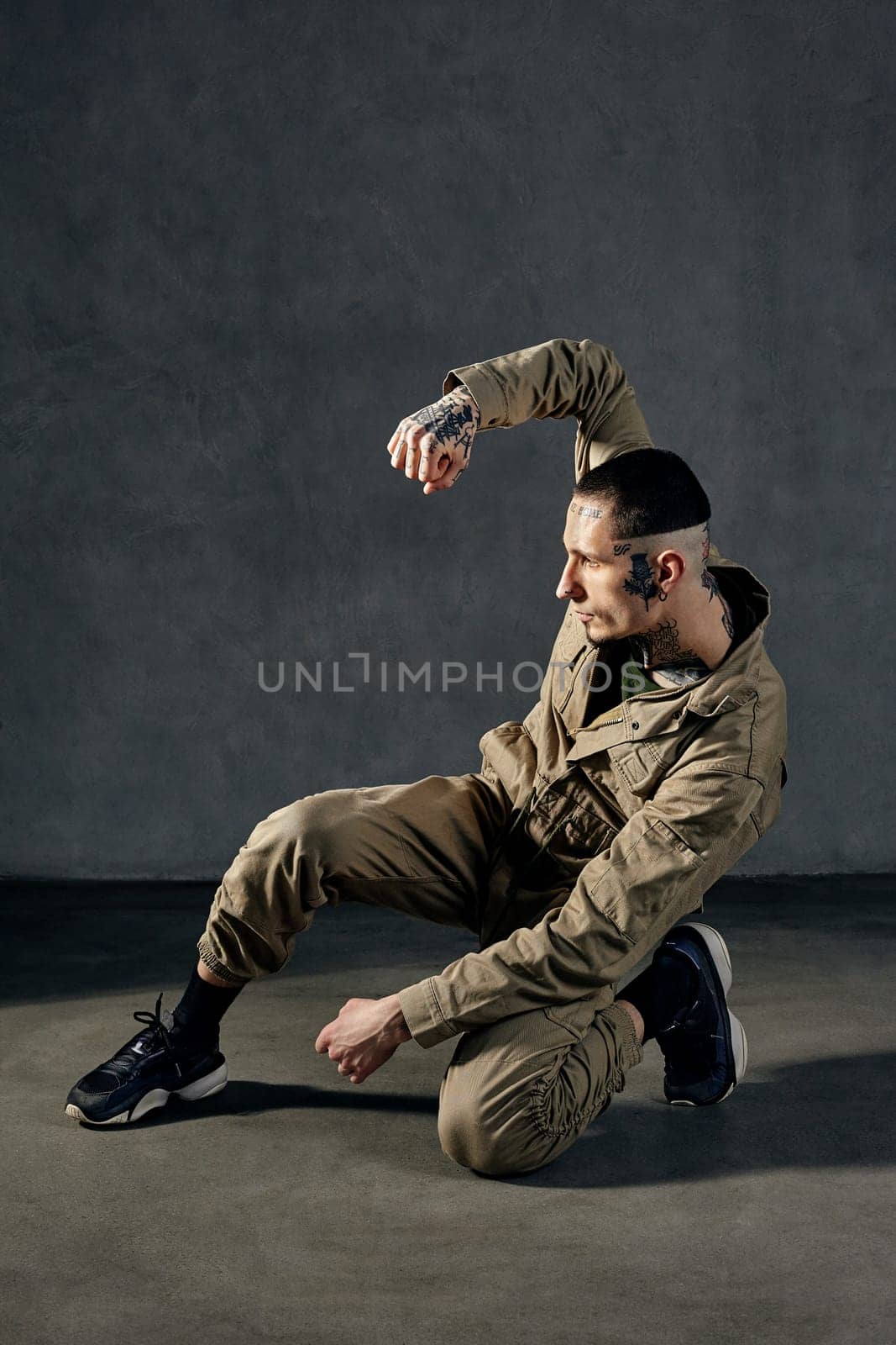 Young athletic man with tattooed body and face, earrings, beard. Dressed in khaki overalls and black sneakers. He is dancing against gray studio background. Dancehall, hip-hop. Full length, copy space