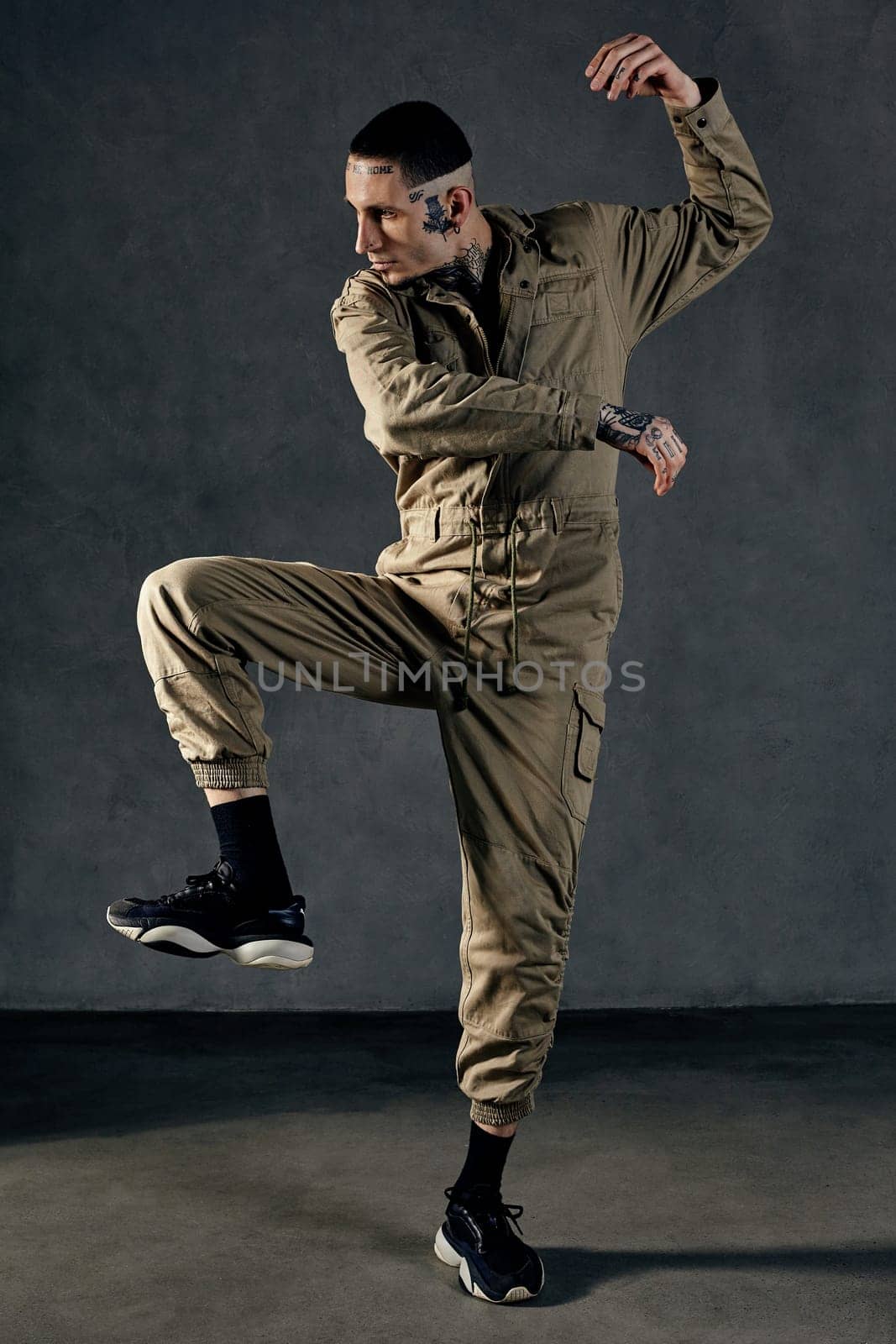 Young athletic male with tattooed body and face, earrings, beard. Dressed in khaki jumpsuit and black sneakers. He dancing against gray studio background. Dancehall, hip-hop. Full length, copy space