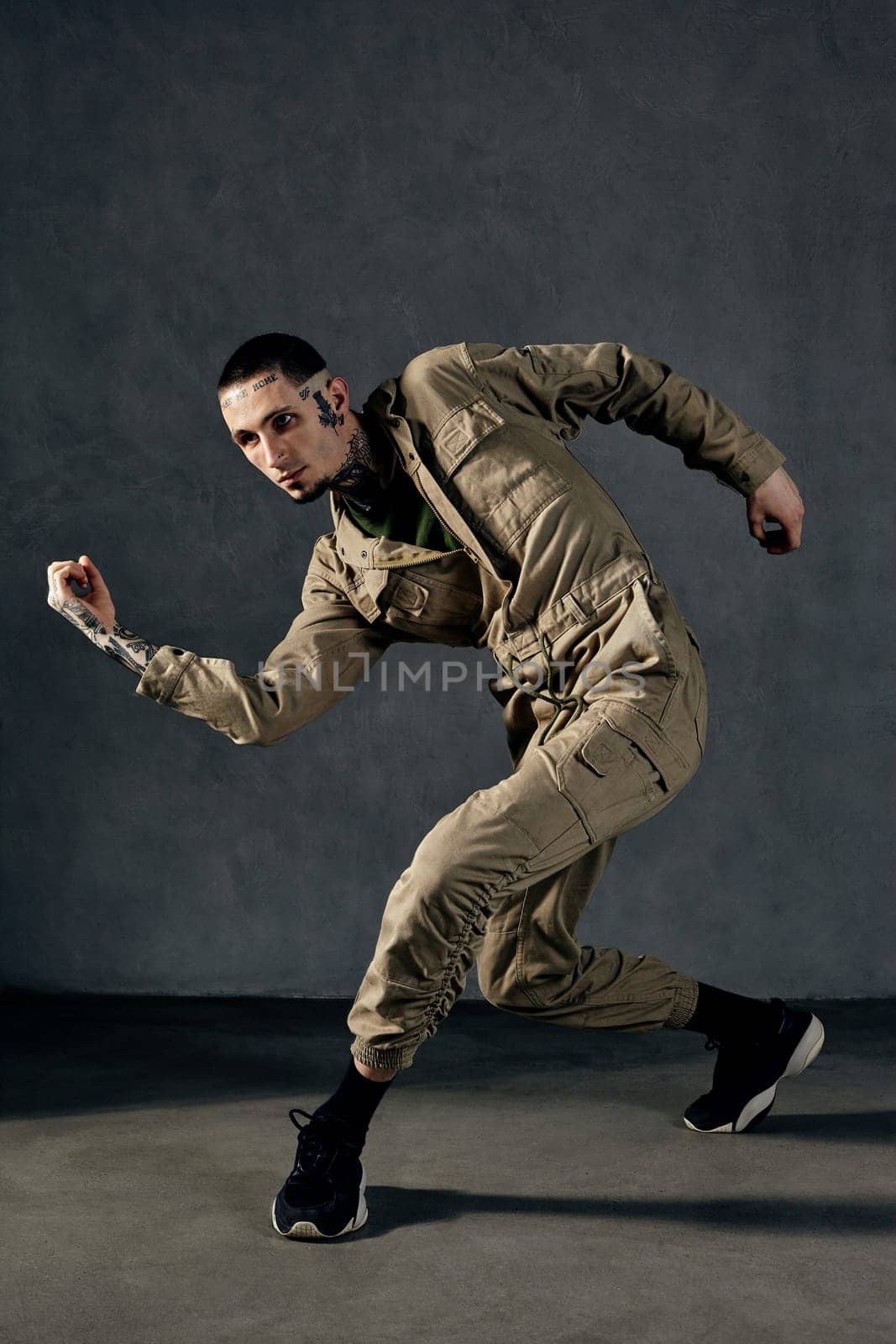 Young athletic performer with tattooed body and face, earrings, beard. Dressed in khaki overalls and black sneakers. He is dancing against gray background. Dancehall, hip-hop. Full length, copy space