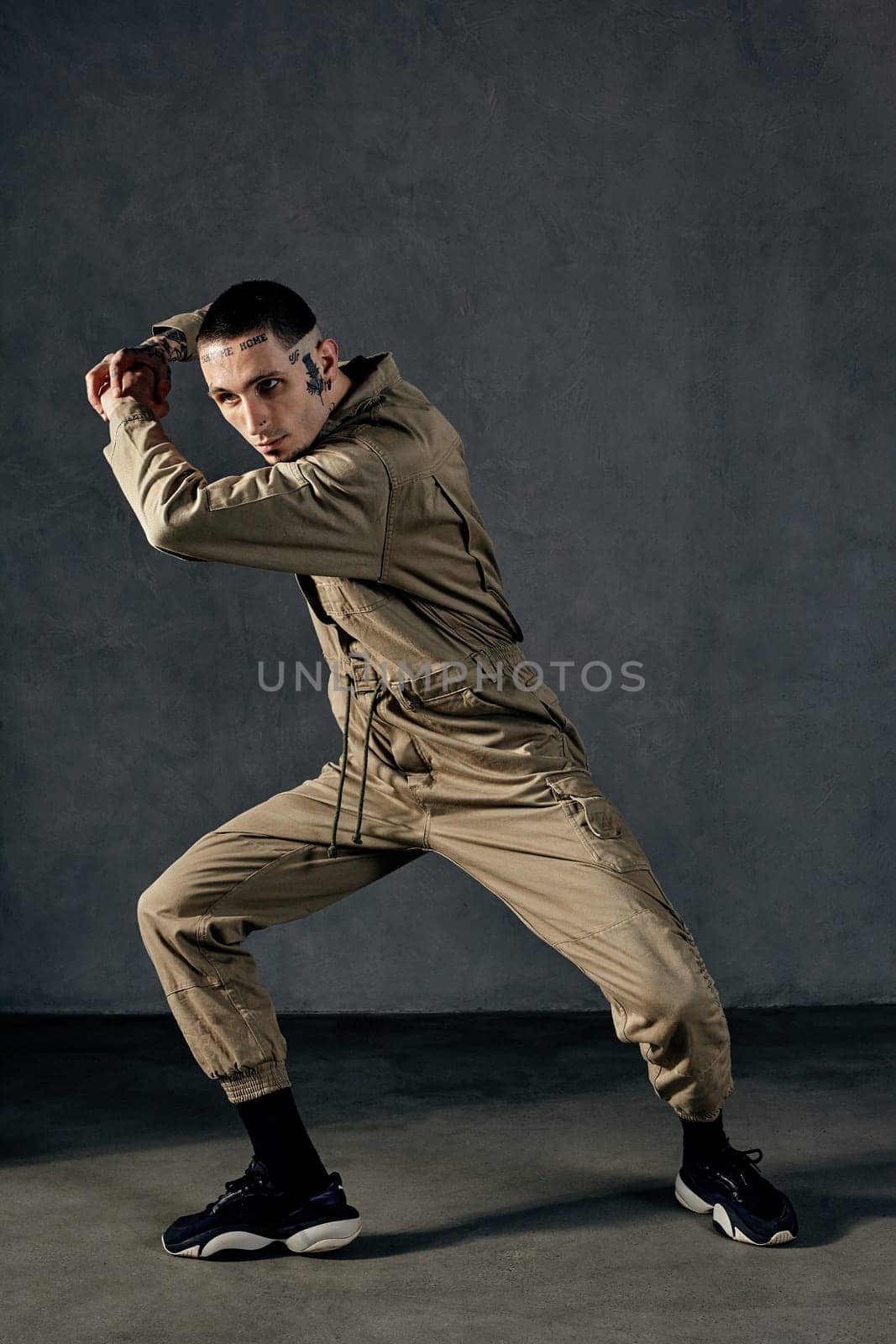 Good-looking male with tattooed body and face, earrings, beard. Dressed in khaki jumpsuit and black sneakers. He is dancing against gray studio background. Dancehall, hip-hop. Full length, copy space