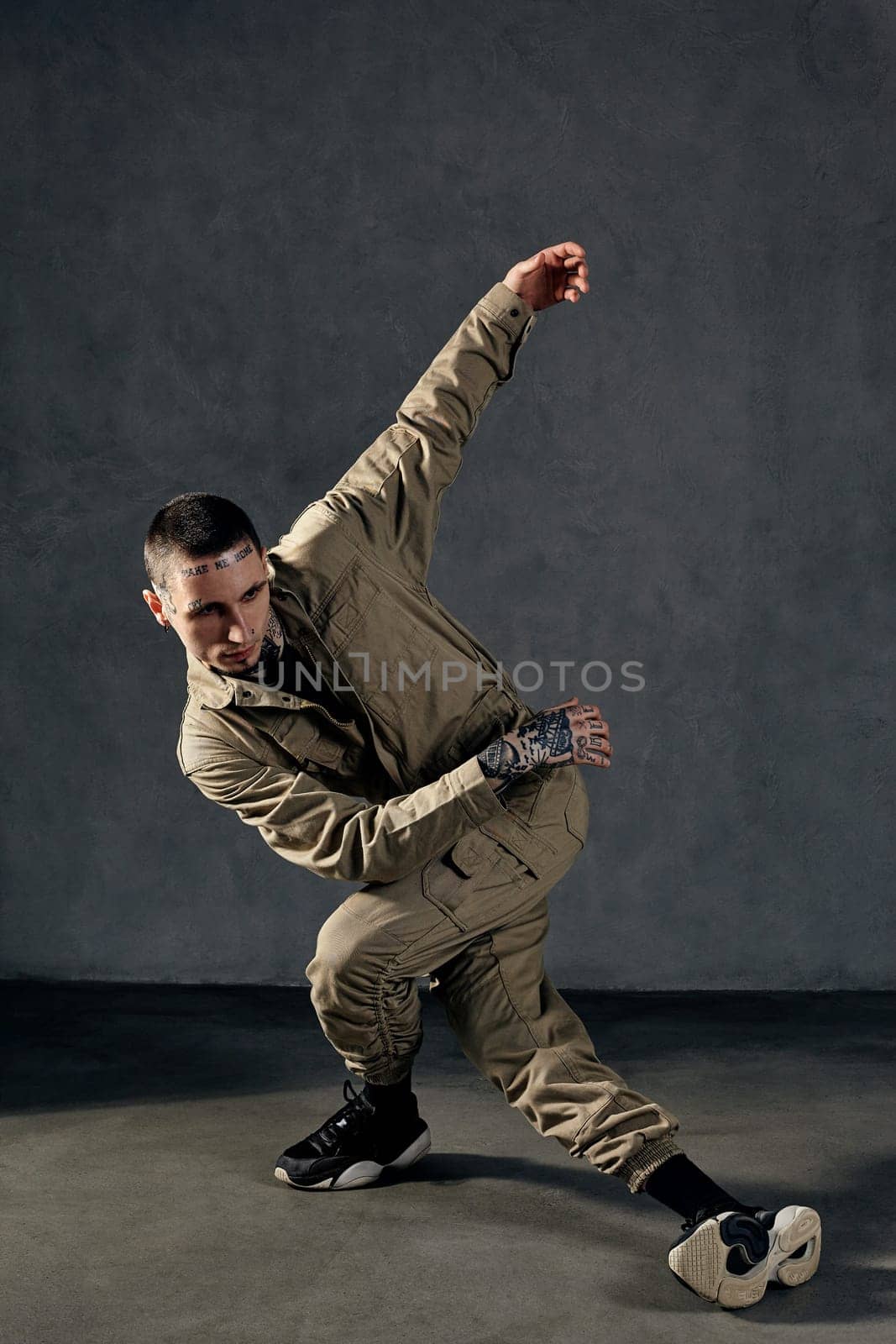 Good-looking fellow with tattooed body and face, earrings, beard. Dressed in khaki jumpsuit and black sneakers. He dancing against gray studio background. Dancehall, hip-hop. Full length, copy space