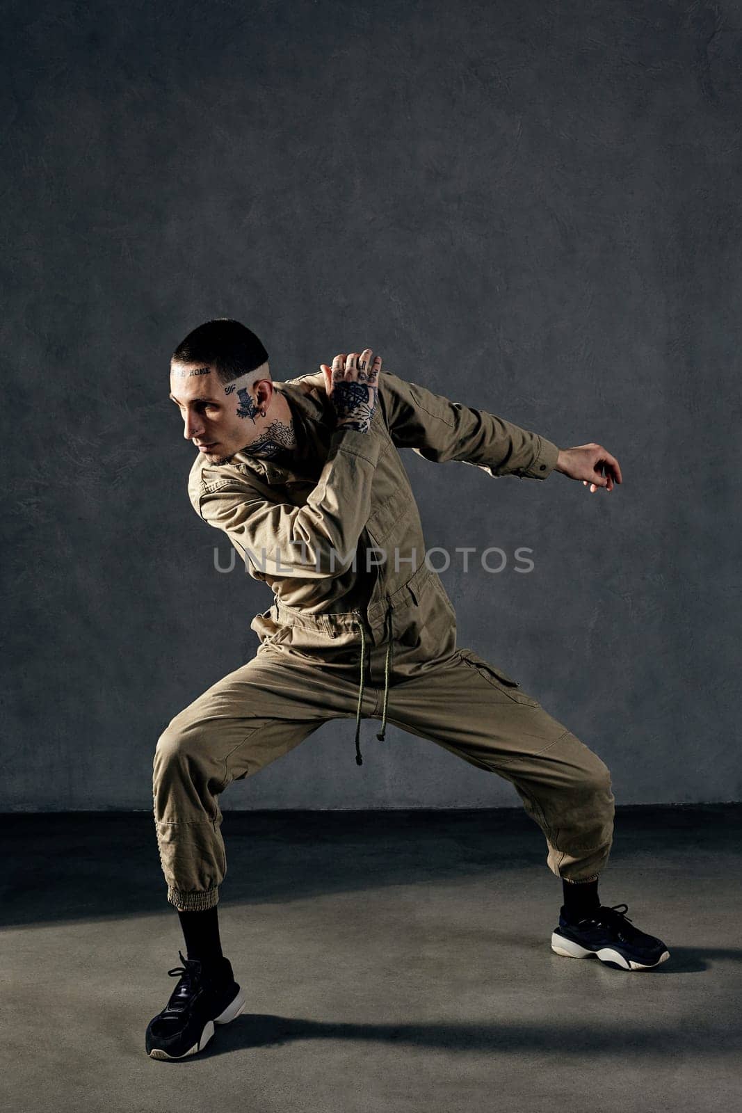 Stately man with tattooed body and face, earrings, beard. Dressed in khaki overalls and black sneakers. Dancing on gray background. Dancehall, hip-hop by nazarovsergey