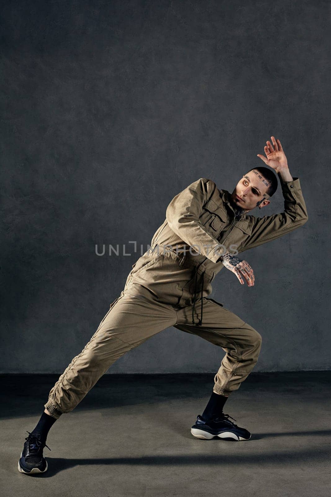 Good-looking performer with tattooed body, earrings, beard. Dressed in khaki overalls, black sneakers. Dancing on gray background. Dancehall, hip-hop by nazarovsergey