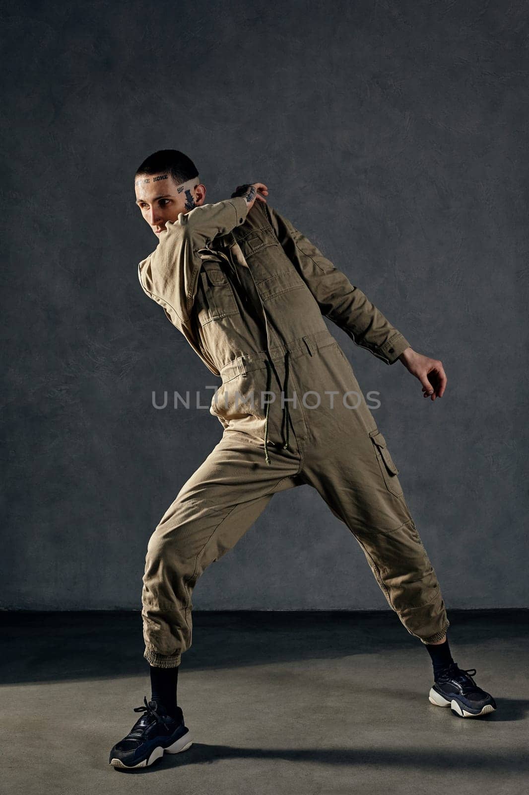 Young stately male with tattooed body and face, earrings, beard. Dressed in khaki jumpsuit and black sneakers. He is dancing against gray studio background. Dancehall, hip-hop. Full length, copy space