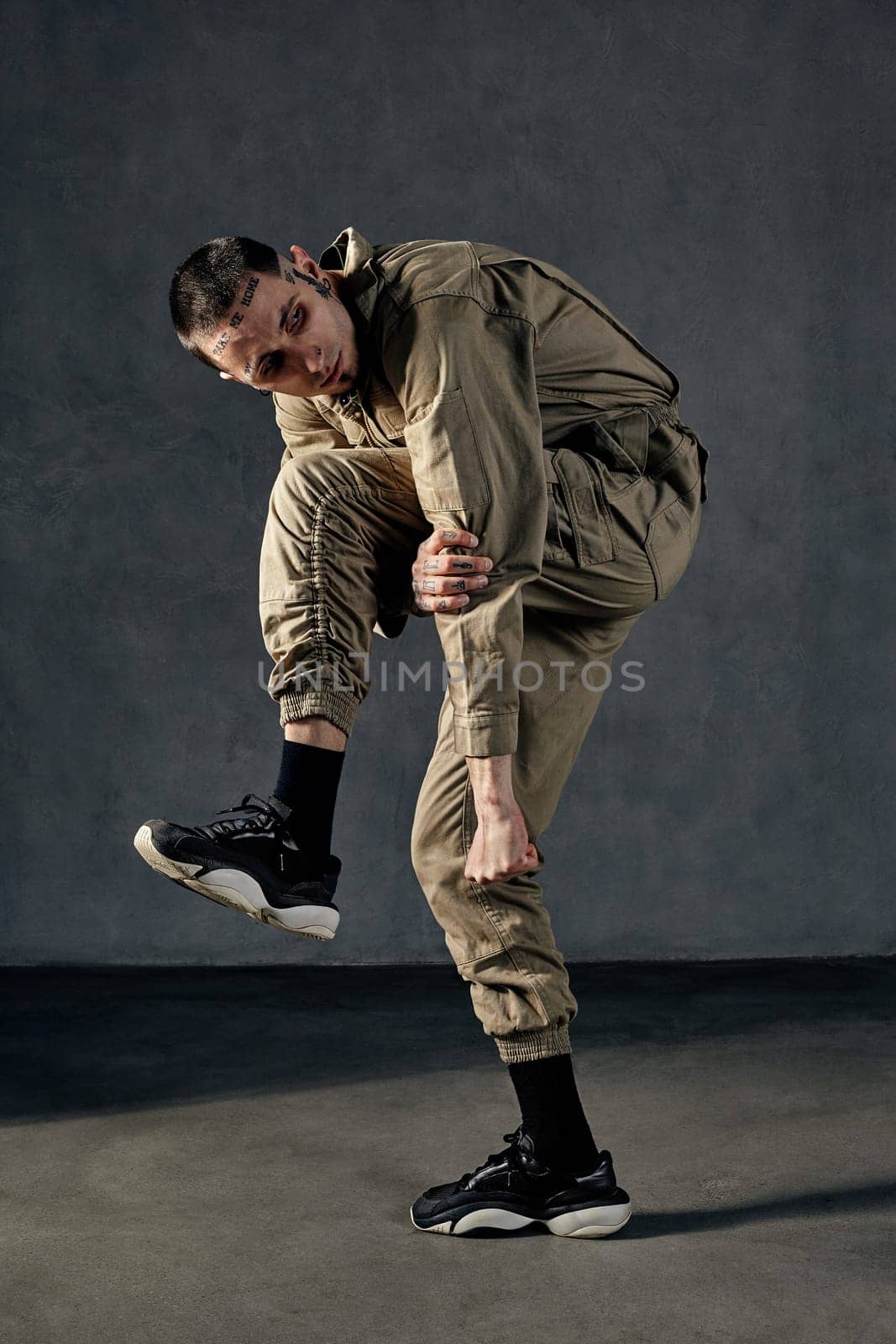 Young strong male with tattooed body and face, earrings, beard. Dressed in khaki jumpsuit and black sneakers. He is dancing against gray studio background. Dancehall, hip-hop. Full length, copy space