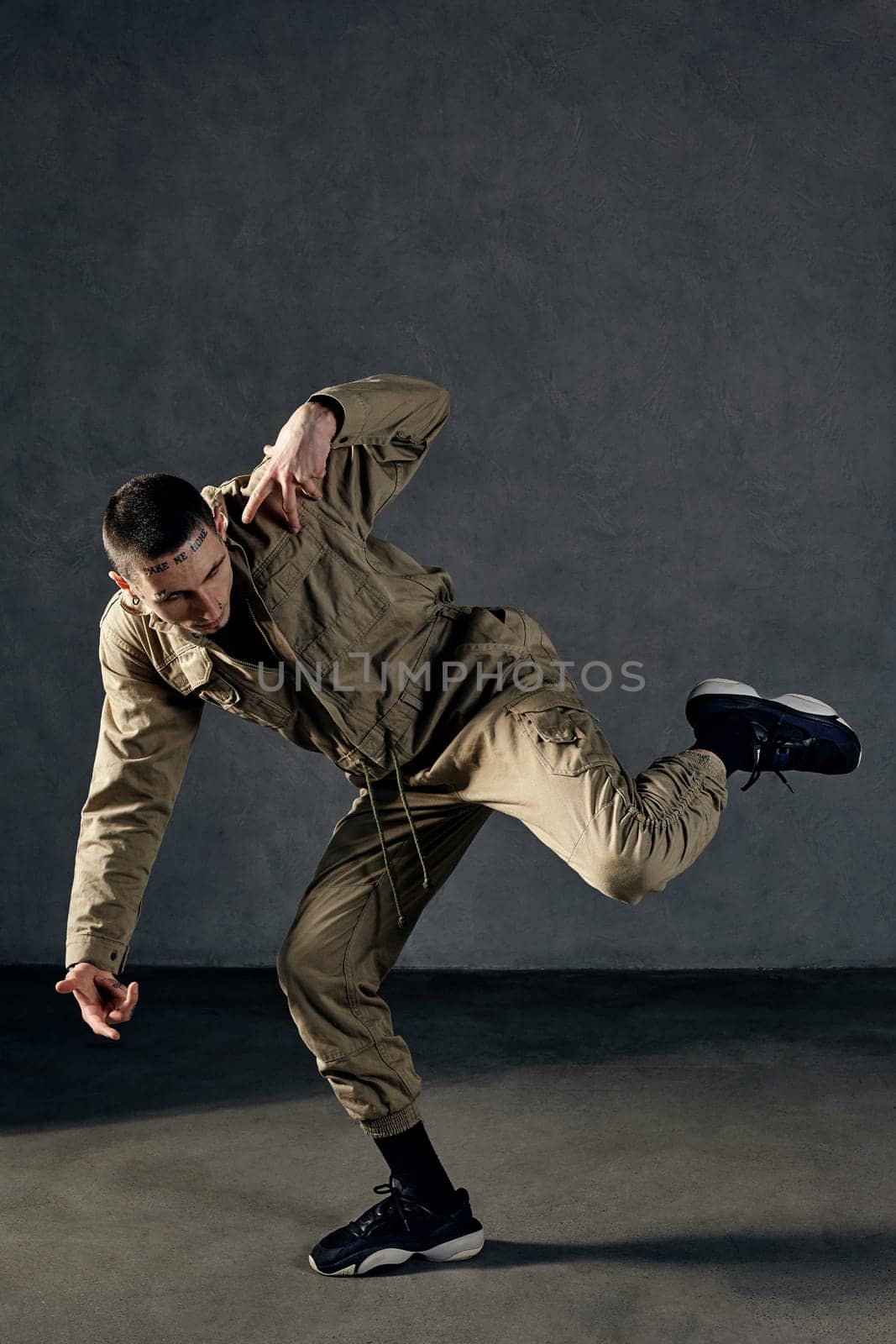 Young strong fellow with tattooed body and face, earrings, beard. Dressed in khaki jumpsuit and black sneakers. He dancing against gray studio background. Dancehall, hip-hop. Full length, copy space