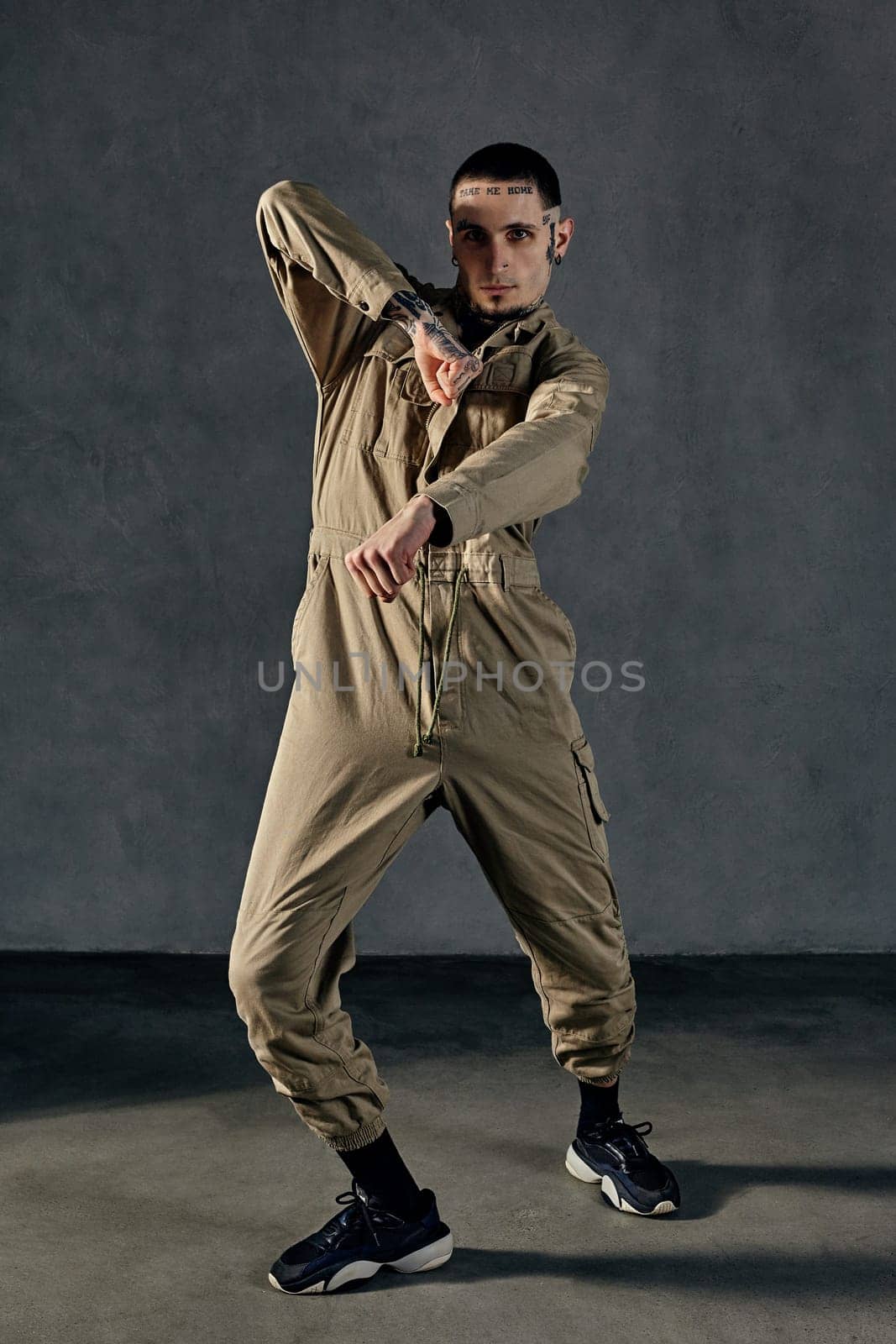 Young active man with tattooed body and face, earrings, beard. Dressed in khaki overalls and black sneakers. He is dancing against gray studio background. Dancehall, hip-hop. Full length, copy space