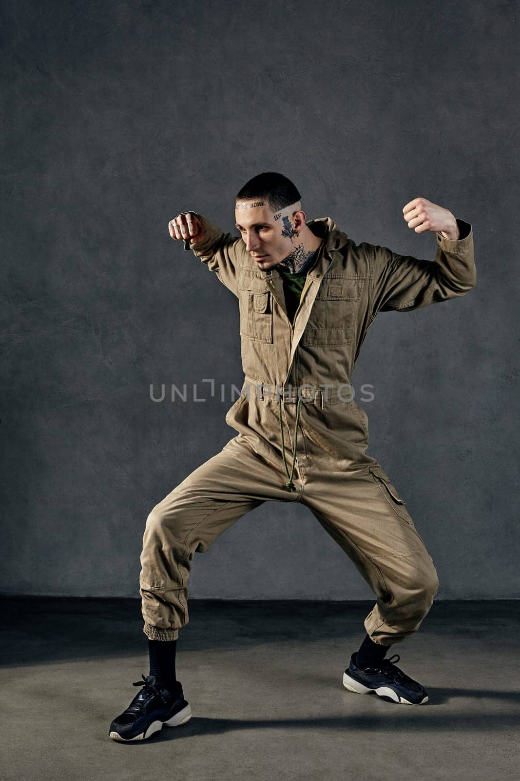 Active male with tattooed body and face, earrings, beard. Dressed in khaki jumpsuit and black sneakers. Dancing on gray background. Dancehall, hip-hop by nazarovsergey