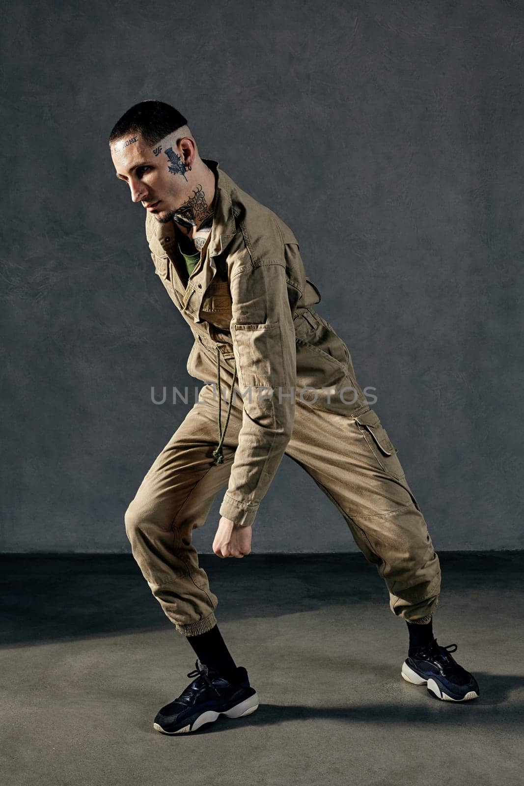 Young active fellow with tattooed body and face, earrings, beard. Dressed in khaki jumpsuit and black sneakers. He dancing against gray studio background. Dancehall, hip-hop. Full length, copy space