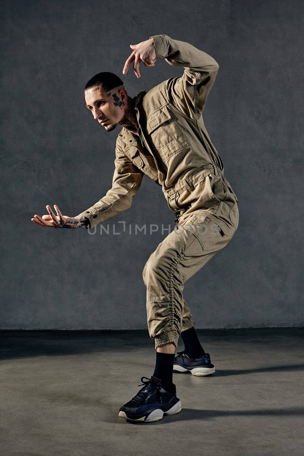 Flexible performer with tattooed body, earrings, beard. Dressed in khaki overalls and black sneakers. Dancing on gray background. Dancehall, hip-hop by nazarovsergey