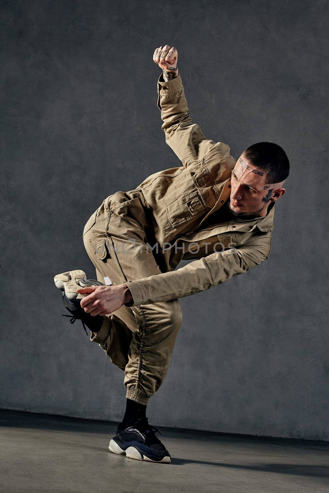 Young attractive male with tattooed body and face, earrings, beard. Dressed in khaki jumpsuit and black sneakers. He dancing against gray studio background. Dancehall, hip-hop. Full length, copy space