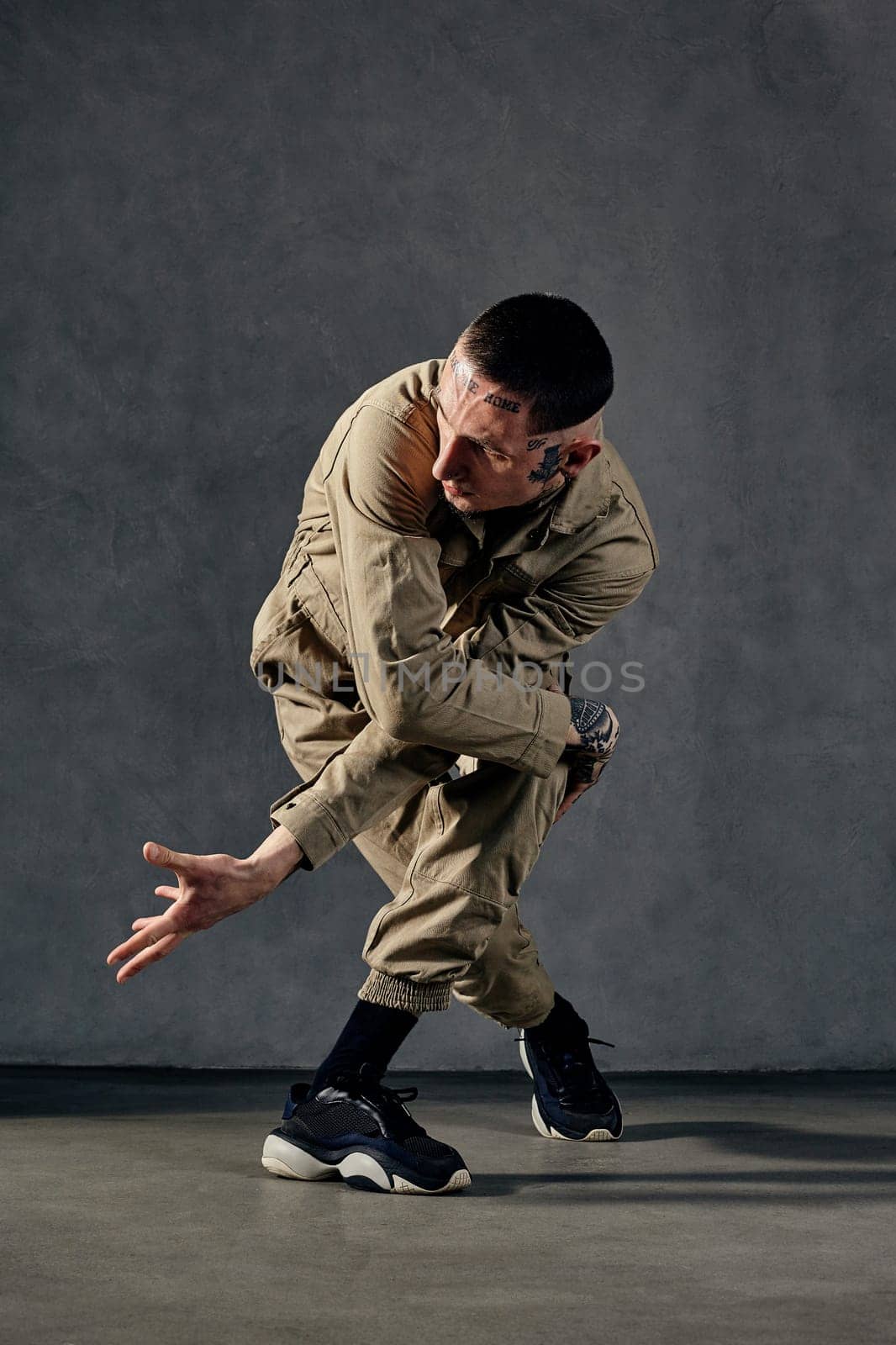Young attractive guy with tattooed body and face, earrings, beard. Dressed in khaki overalls and black sneakers. He dancing against gray studio background. Dancehall, hip-hop. Full length, copy space