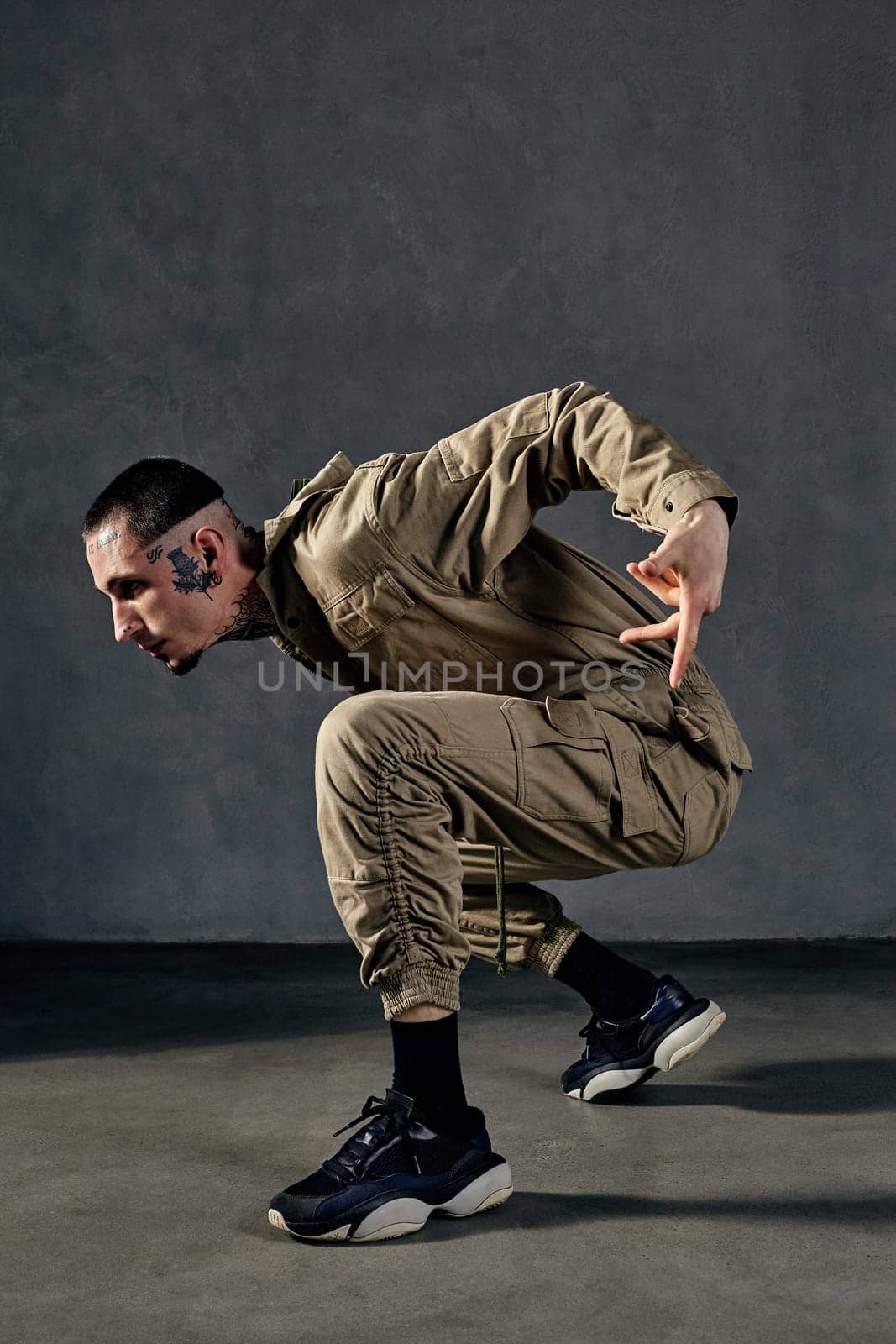 Young attractive performer with tattooed body and face, earrings, beard. Dressed in khaki overalls and black sneakers. He dancing against gray background. Dancehall, hip-hop. Full length, copy space