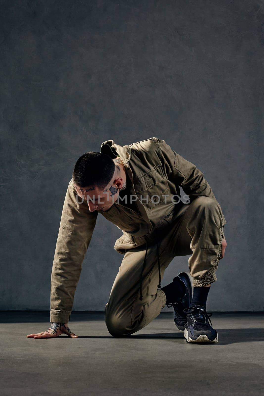 Young unusual male with tattooed body and face, earrings, beard. Dressed in khaki jumpsuit and black sneakers. He is dancing against gray studio background. Dancehall, hip-hop. Full length, copy space